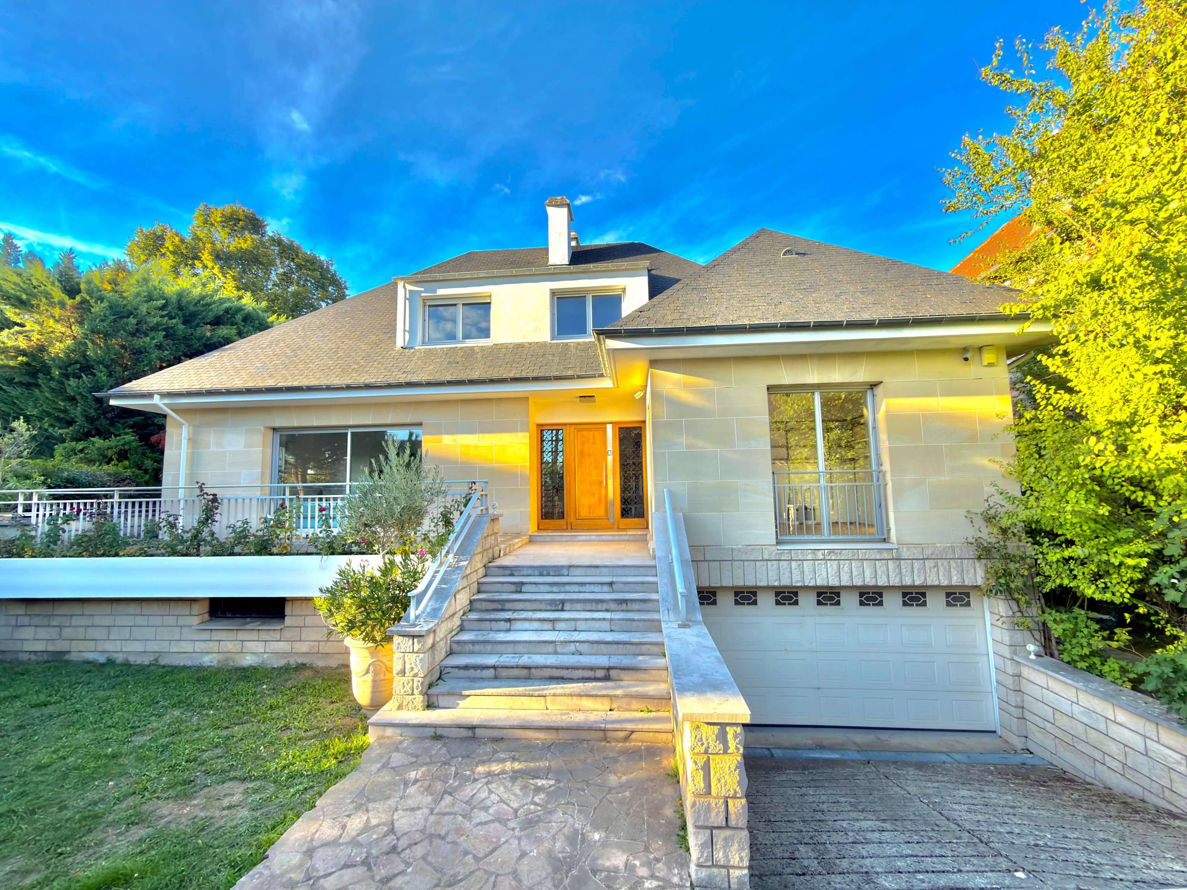 The image shows a large, two-story house with a stone facade and a sloped roof, surrounded by greenery and featuring a driveway leading to a garage.