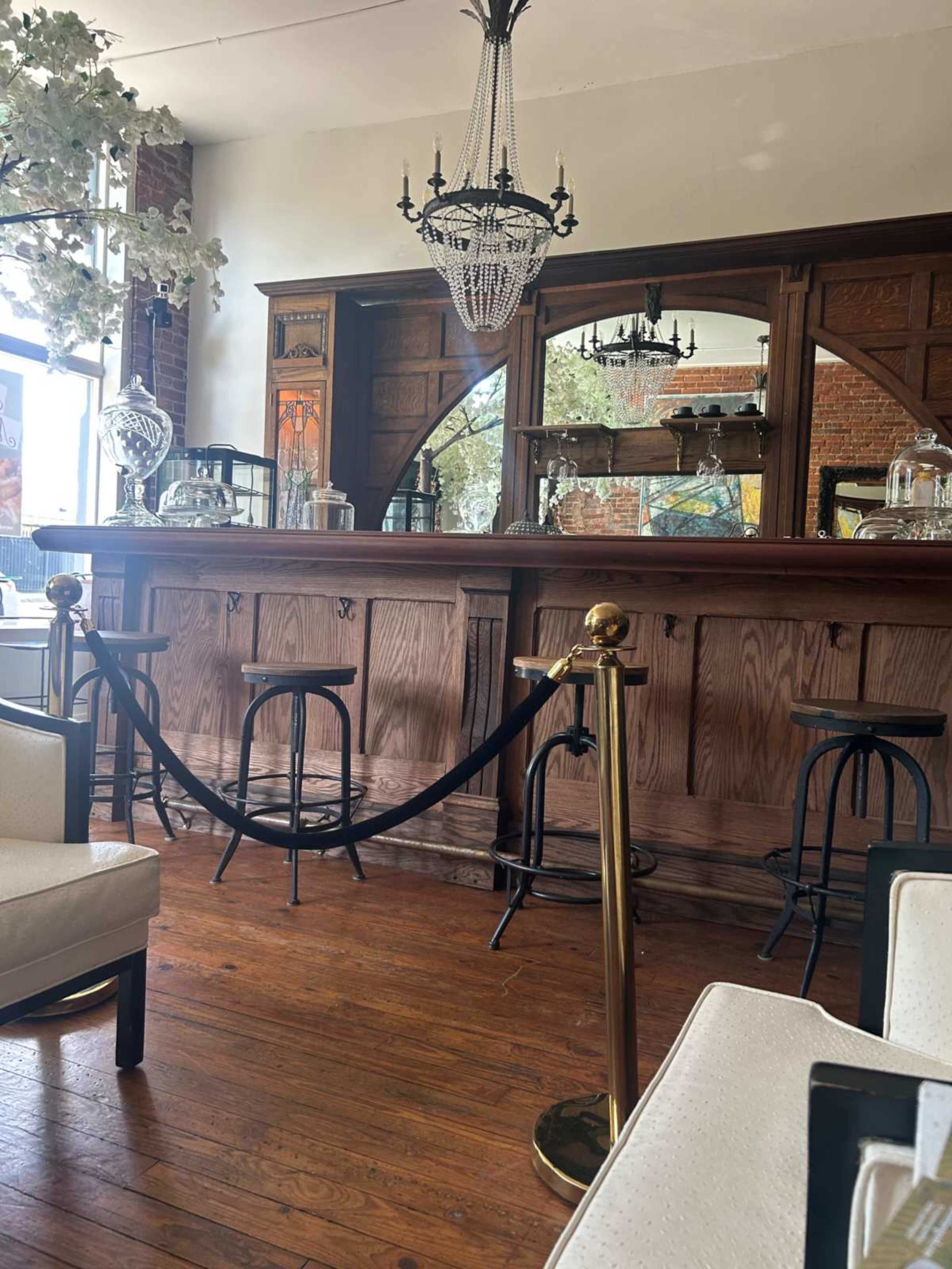 The image shows a wooden bar with high stools and a chandelier, surrounded by a seating area in a well-lit café.