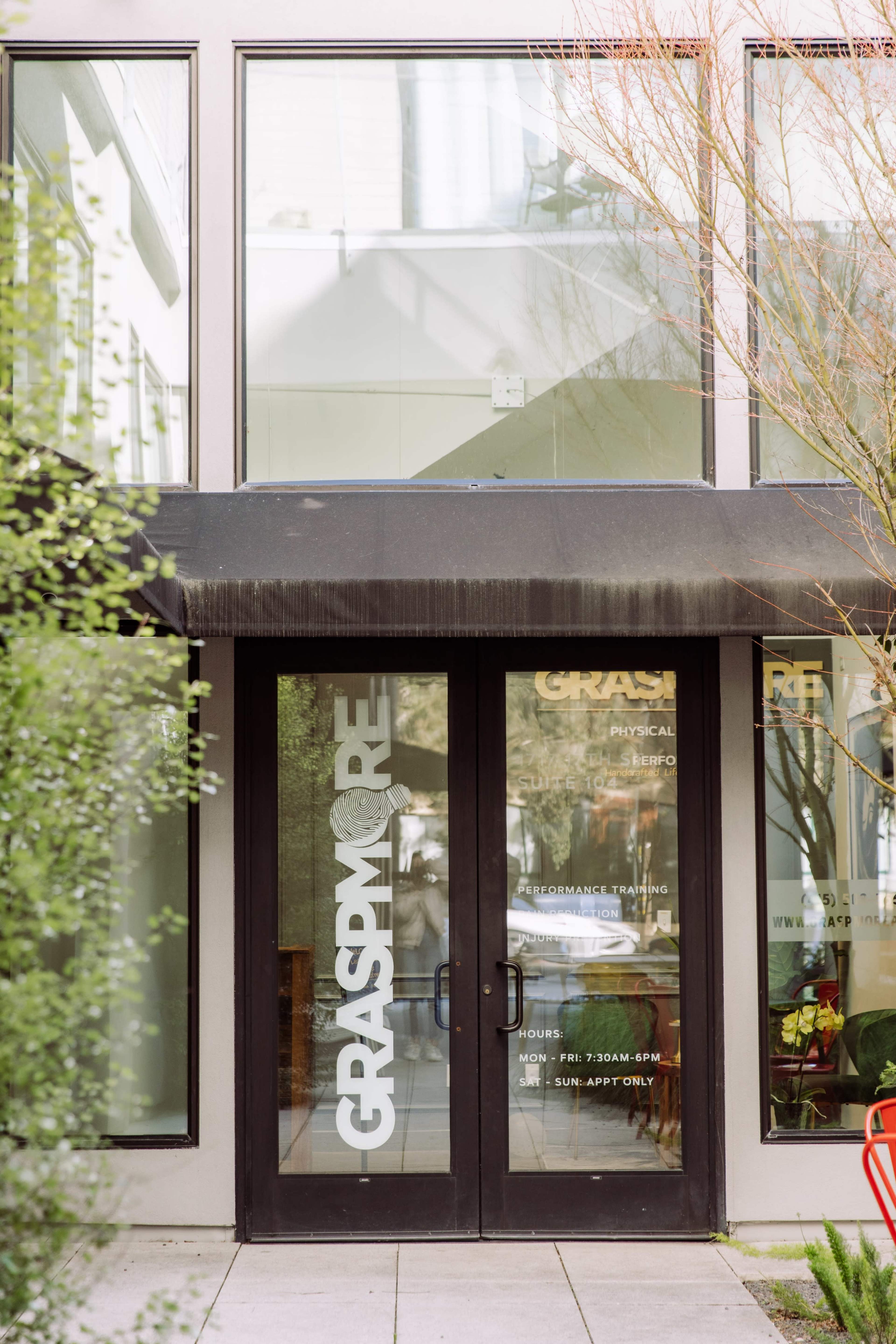 The entrance of a building features large glass windows and a black awning above double doors marked with the name "GRASP MORE."
