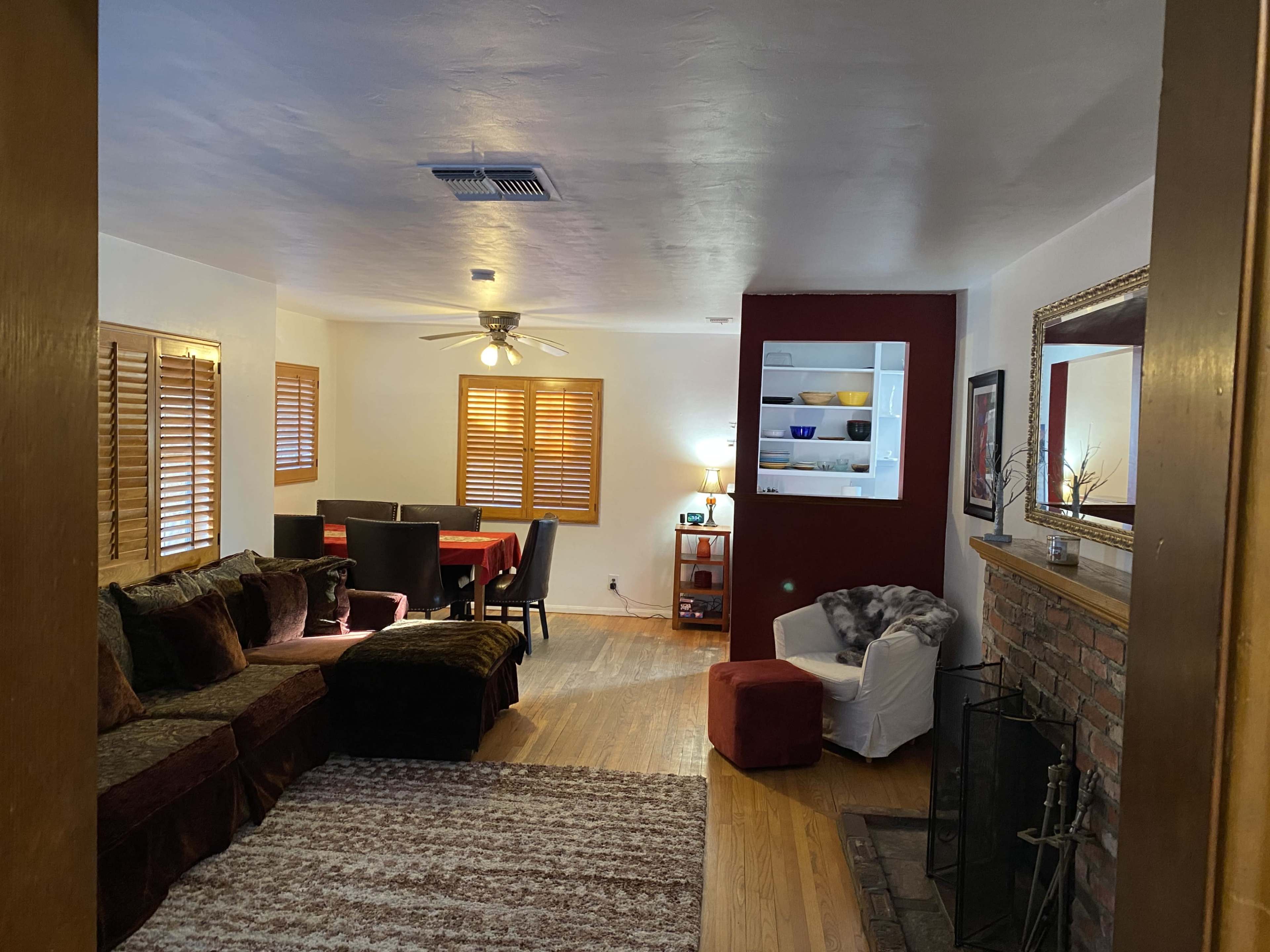 The image shows a cozy living room with a sofa, dining area, and a fireplace, featuring wooden blinds on the windows.