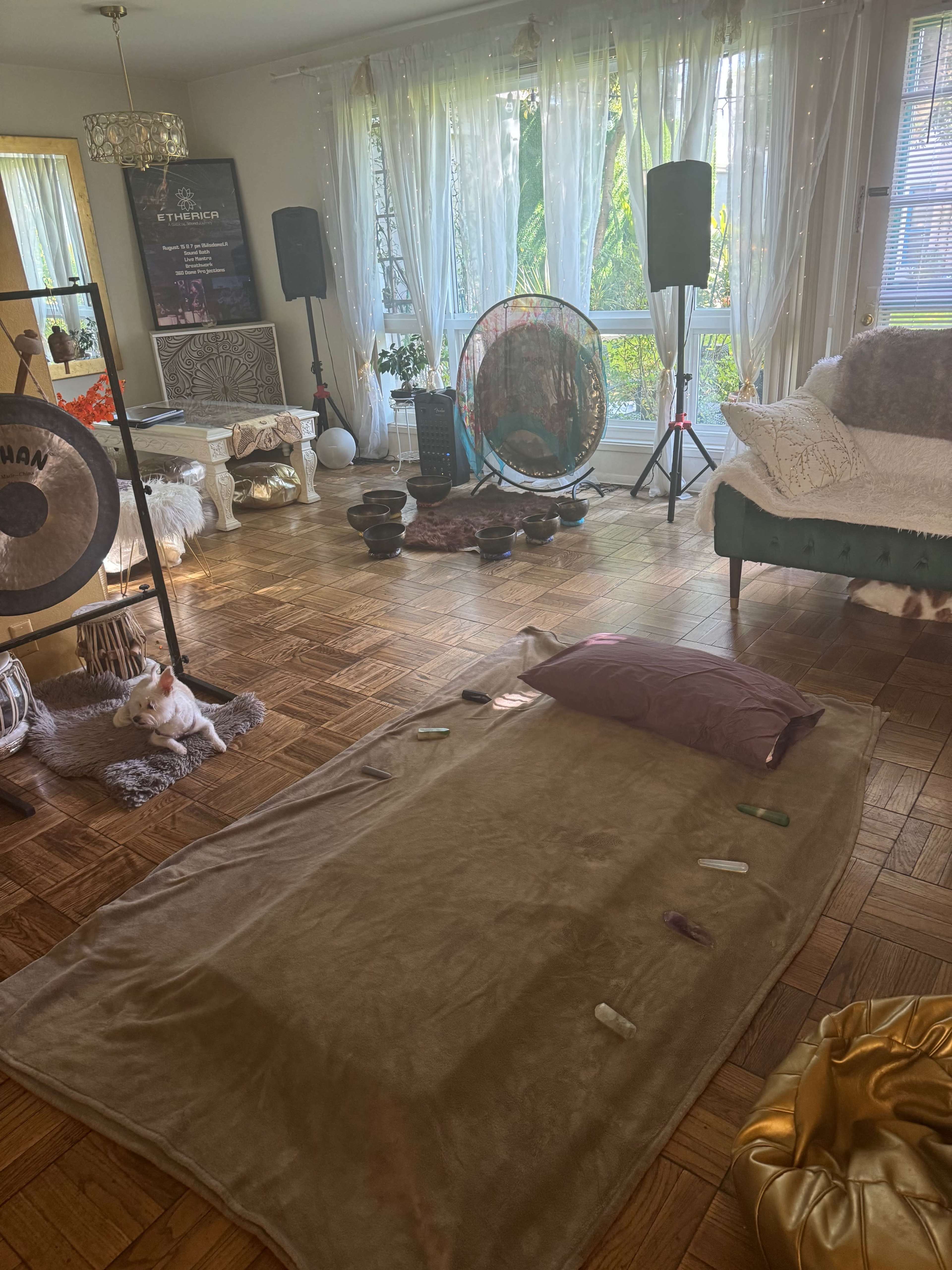 The image shows a spacious room with wooden flooring, decorated with soft furnishings, a large gong, various cushions, and a mat with a pillow on it, set for relaxation or meditation.