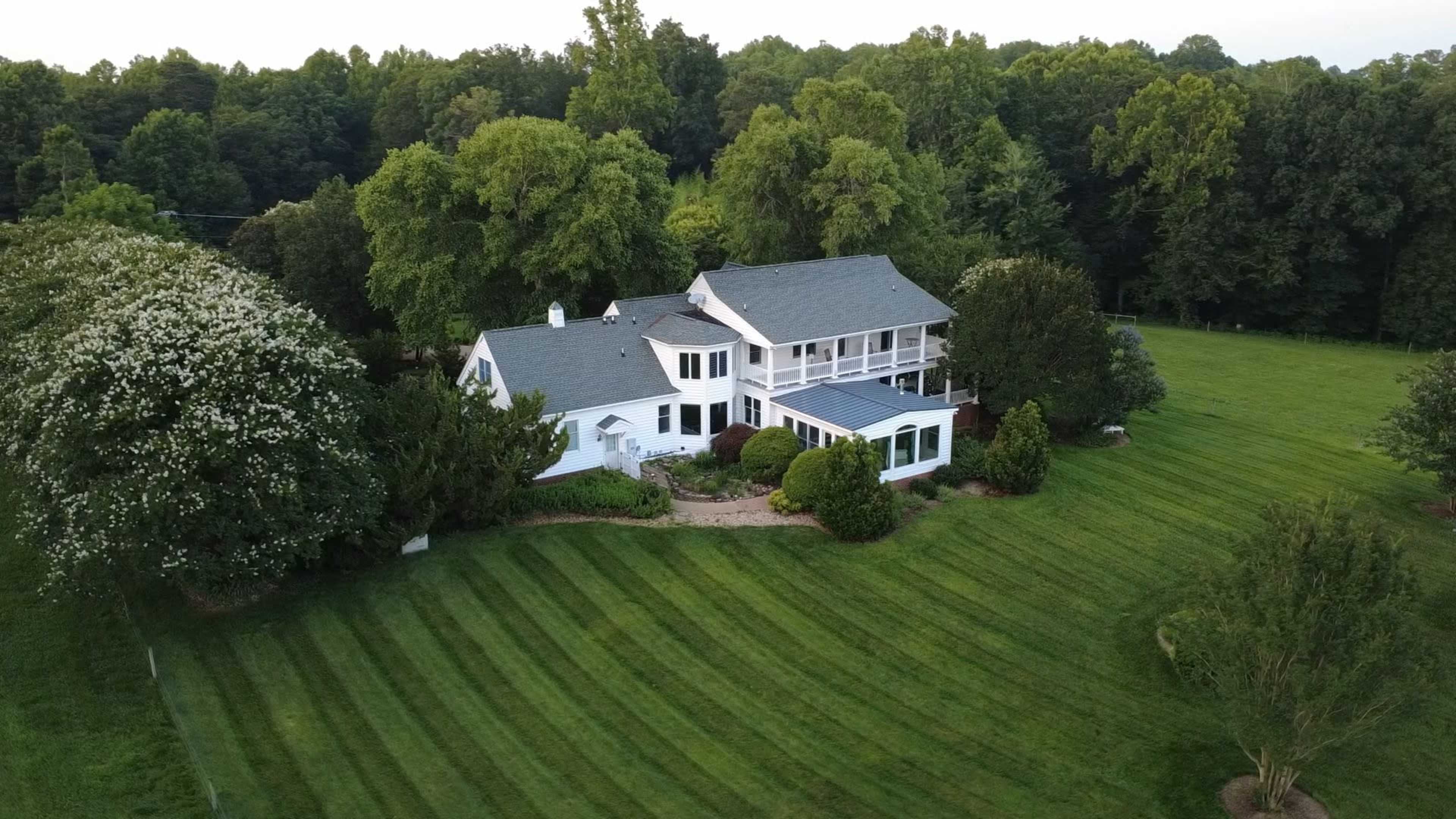 A white house with a large porch is surrounded by neatly striped green lawns and trees.
