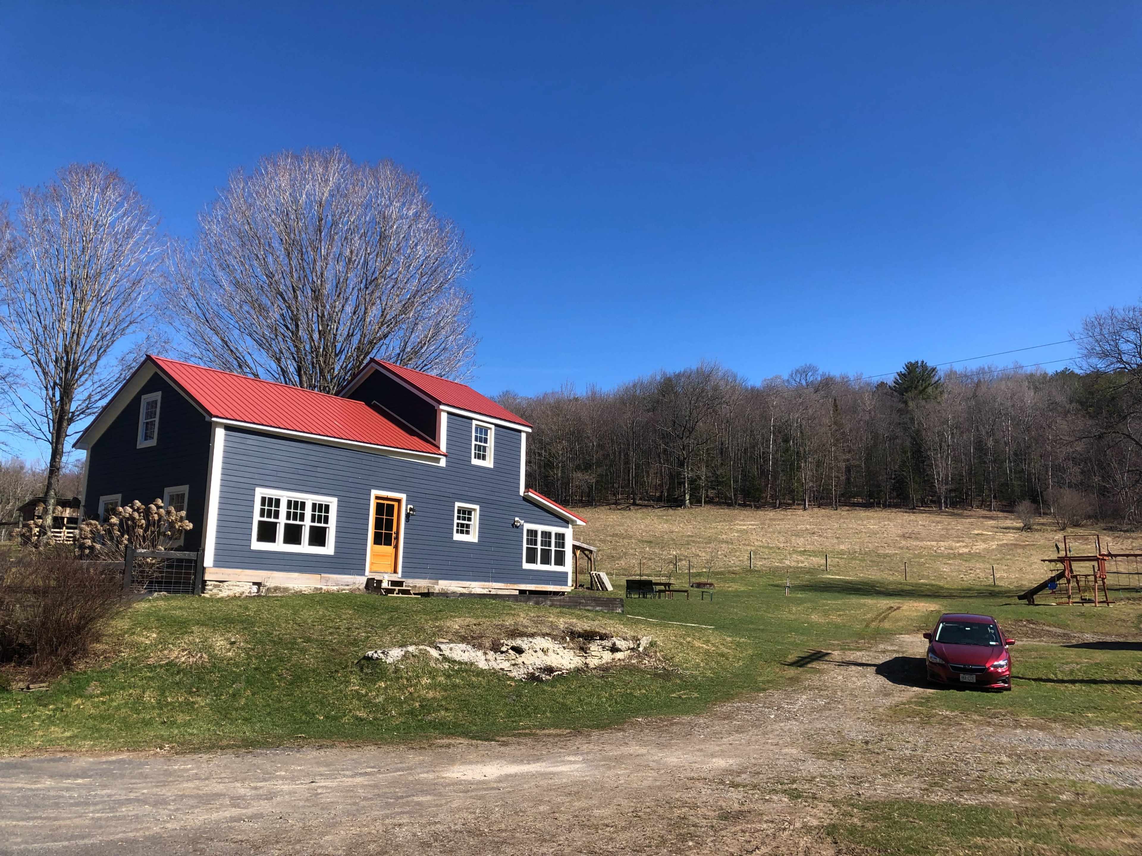 A gray house with a red roof is situated on a grassy area near a dirt driveway, with a car parked nearby and trees in the background.
