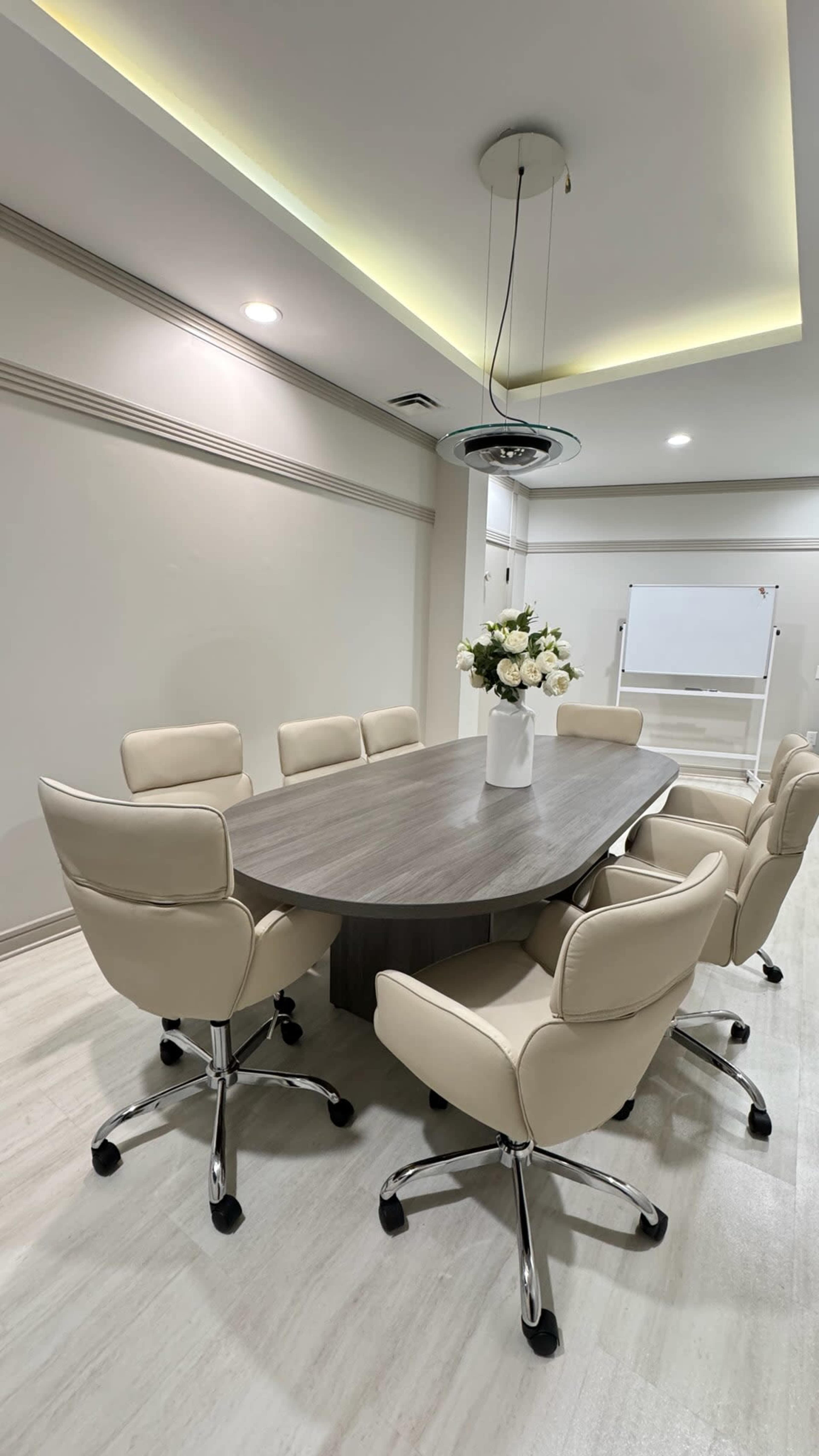 The image shows a modern conference room with a large oval table surrounded by eight beige swivel chairs and a flower arrangement in a vase on the table.