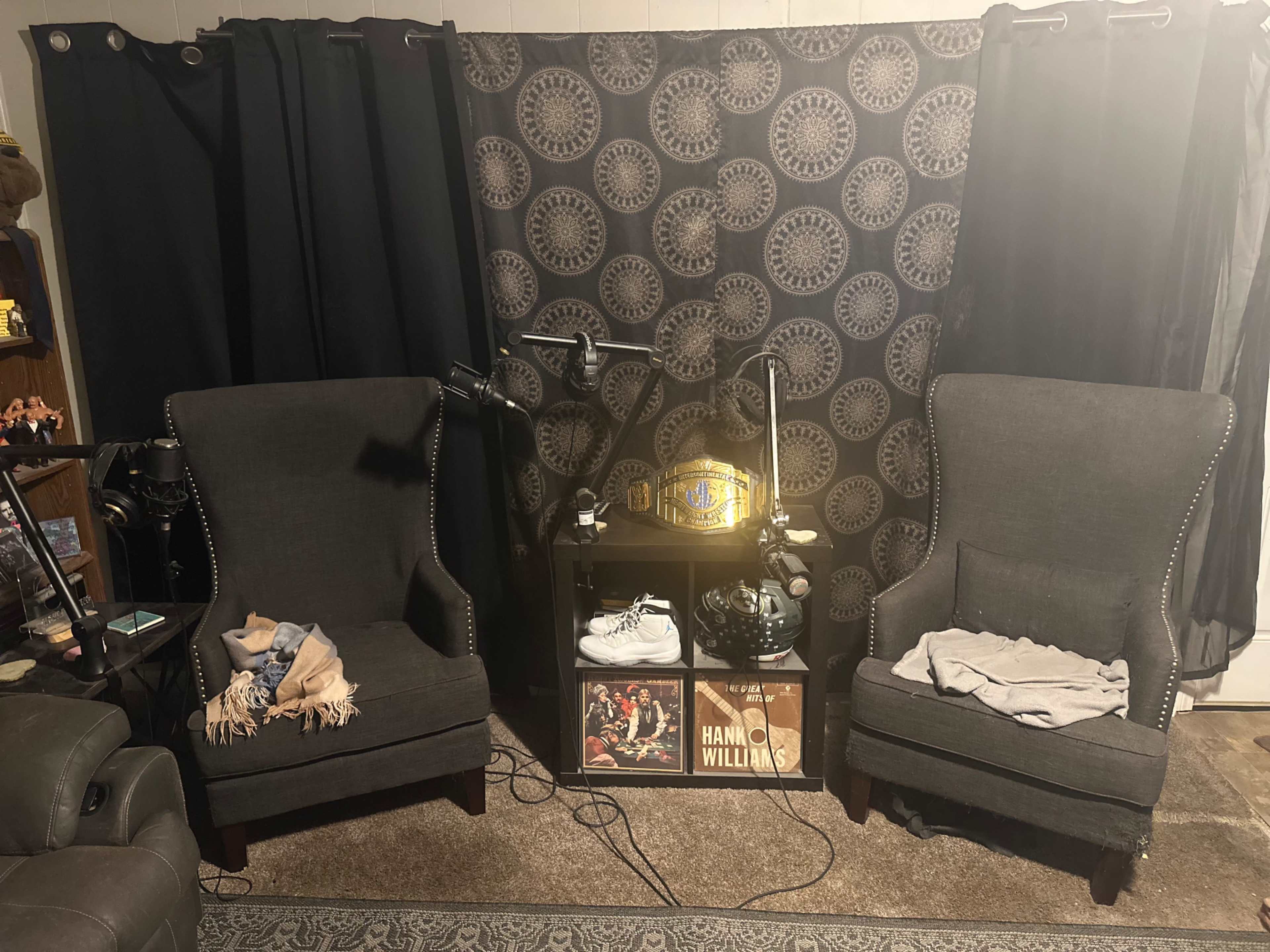 Two dark armchairs facing a small table adorned with a championship belt and some framed records, set against a patterned curtain.