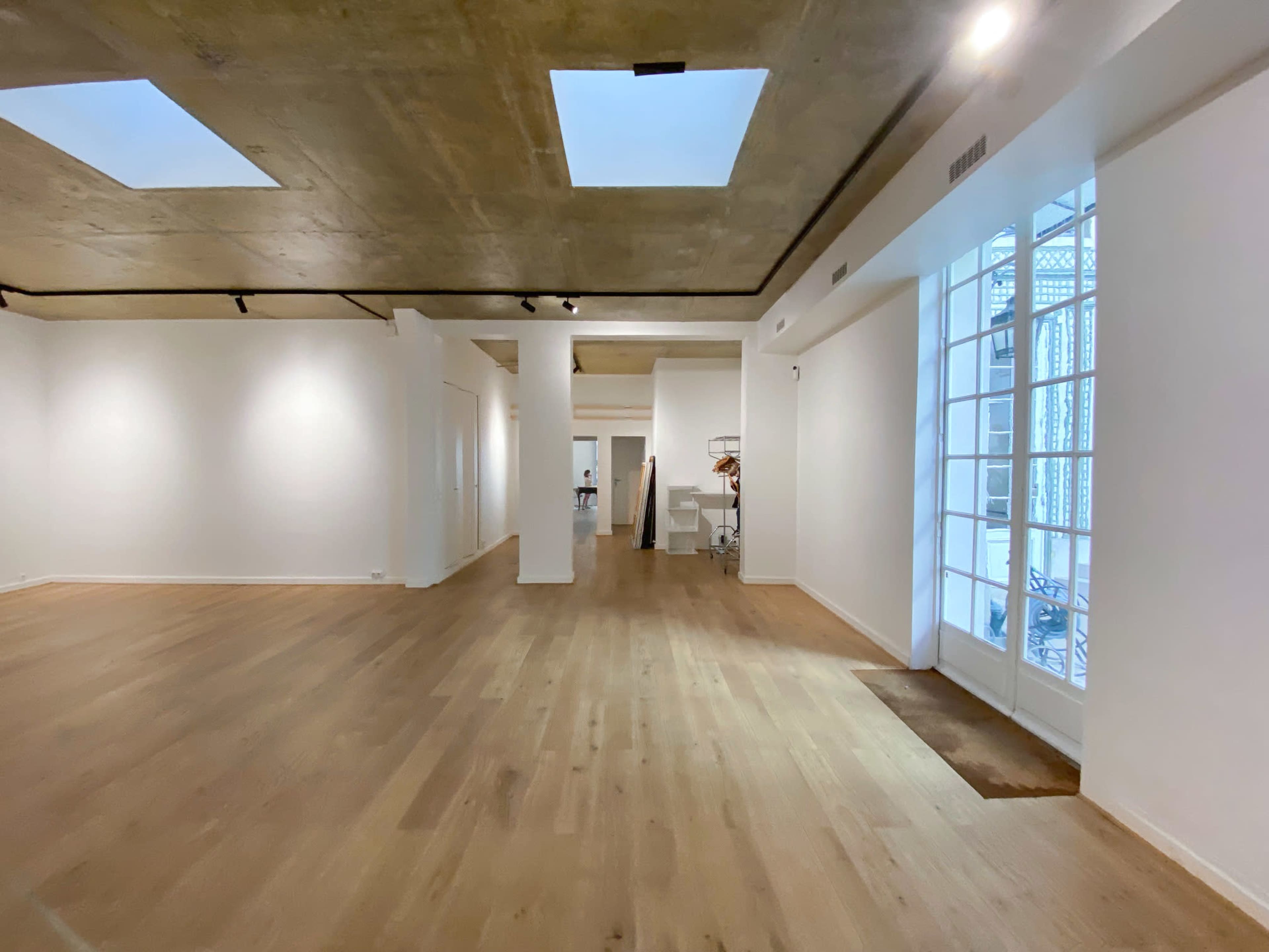 The image shows a spacious, empty gallery with light wood flooring, white walls, and several windows allowing natural light to enter.