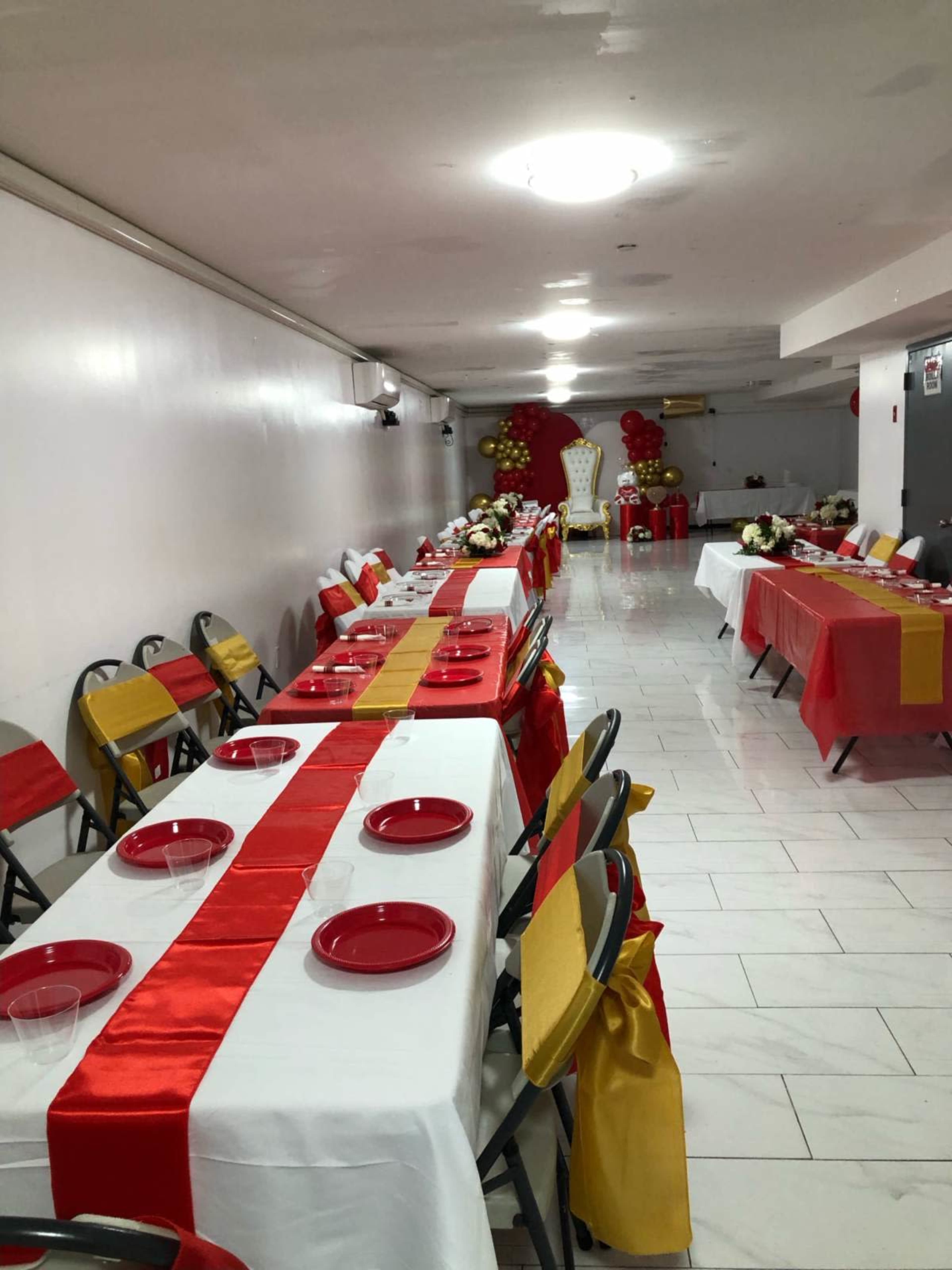 The image shows a banquet hall set up with long tables covered in white tablecloths adorned with red and yellow ribbons, featuring red plates and chairs arranged for an event.