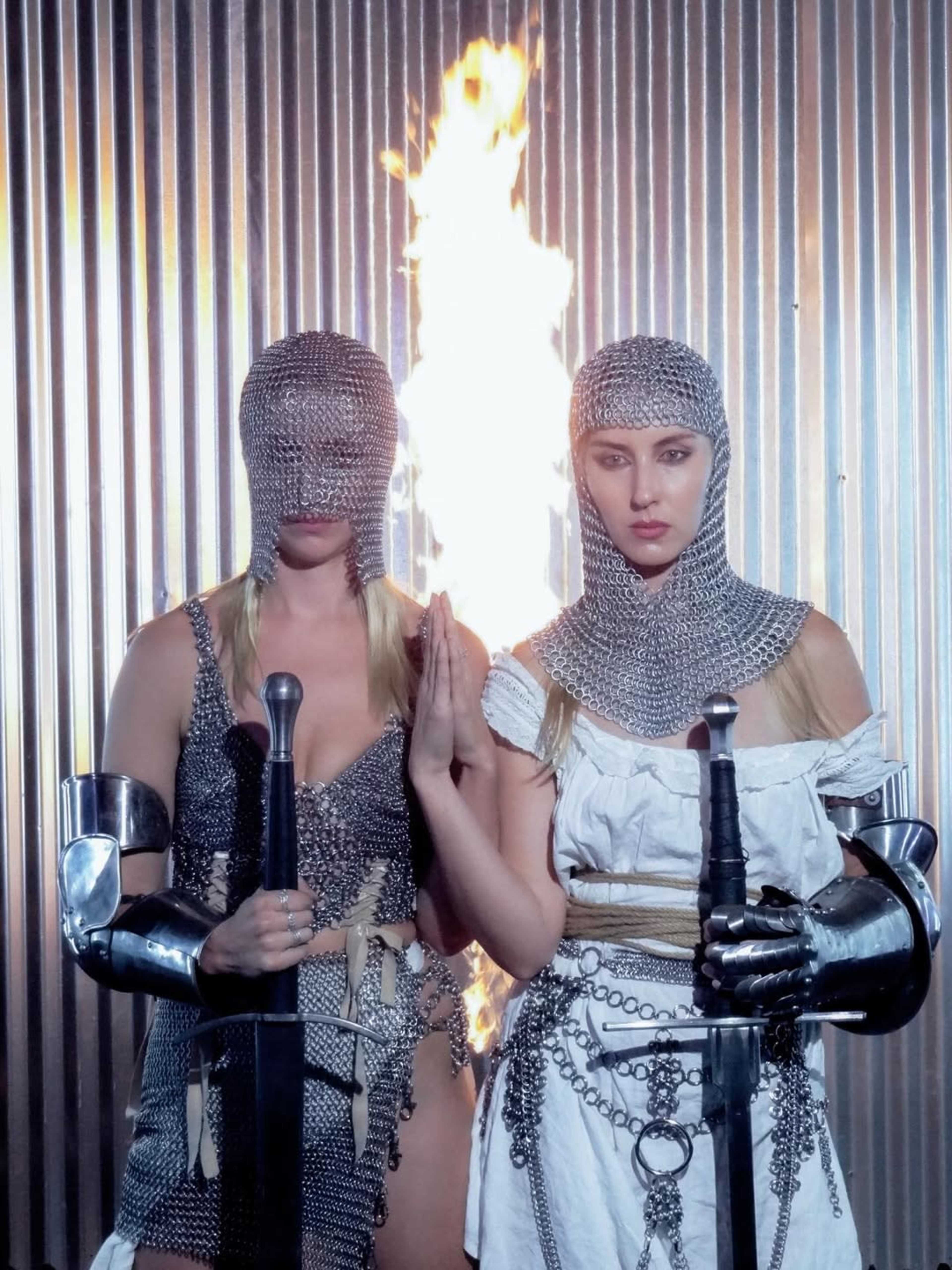 Two women in chainmail armor hold swords in front of a flaming backdrop.
