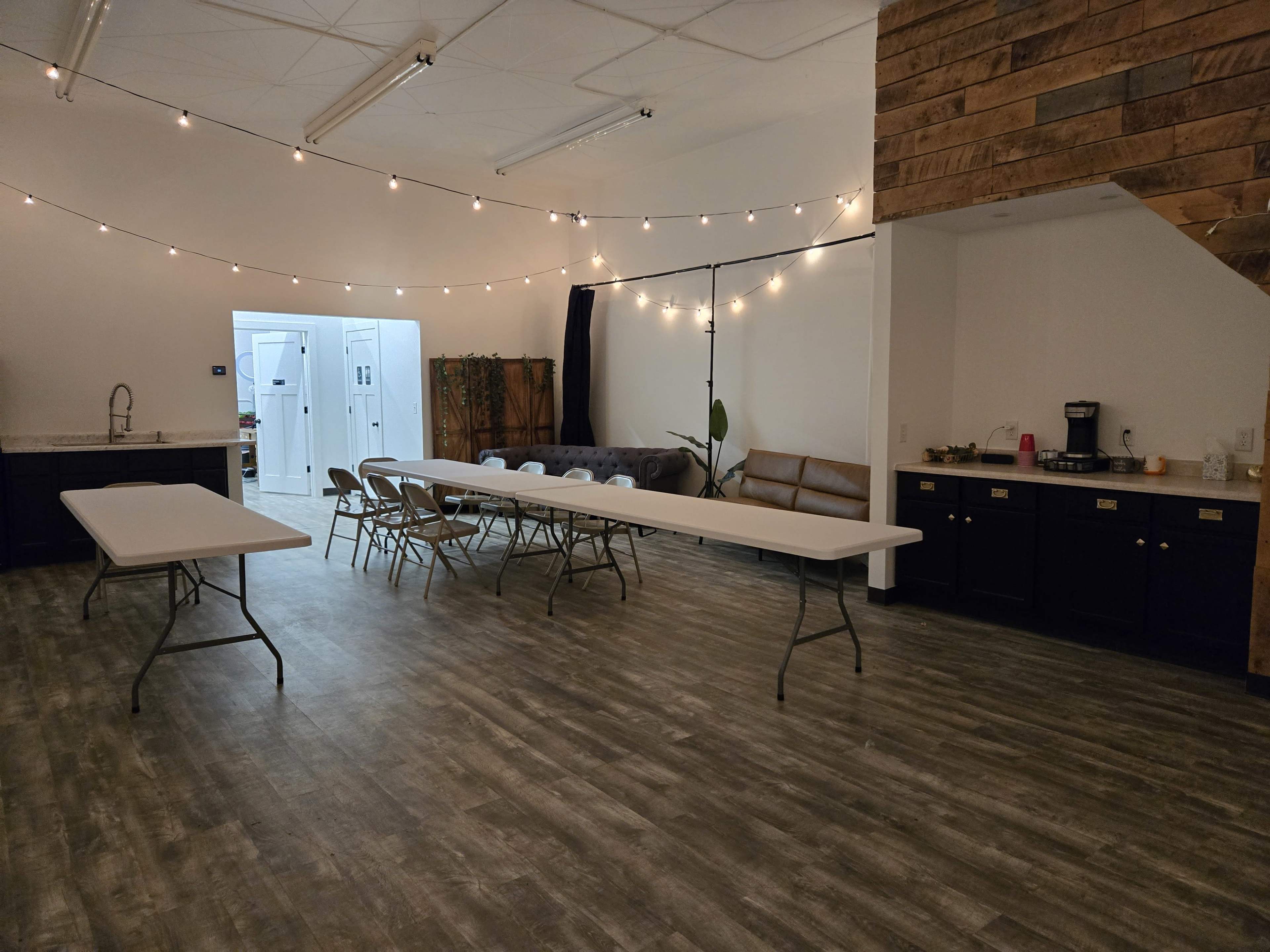 The image shows a spacious room with string lights overhead, featuring a long table with folding chairs, a couch, and a kitchenette area in the background.