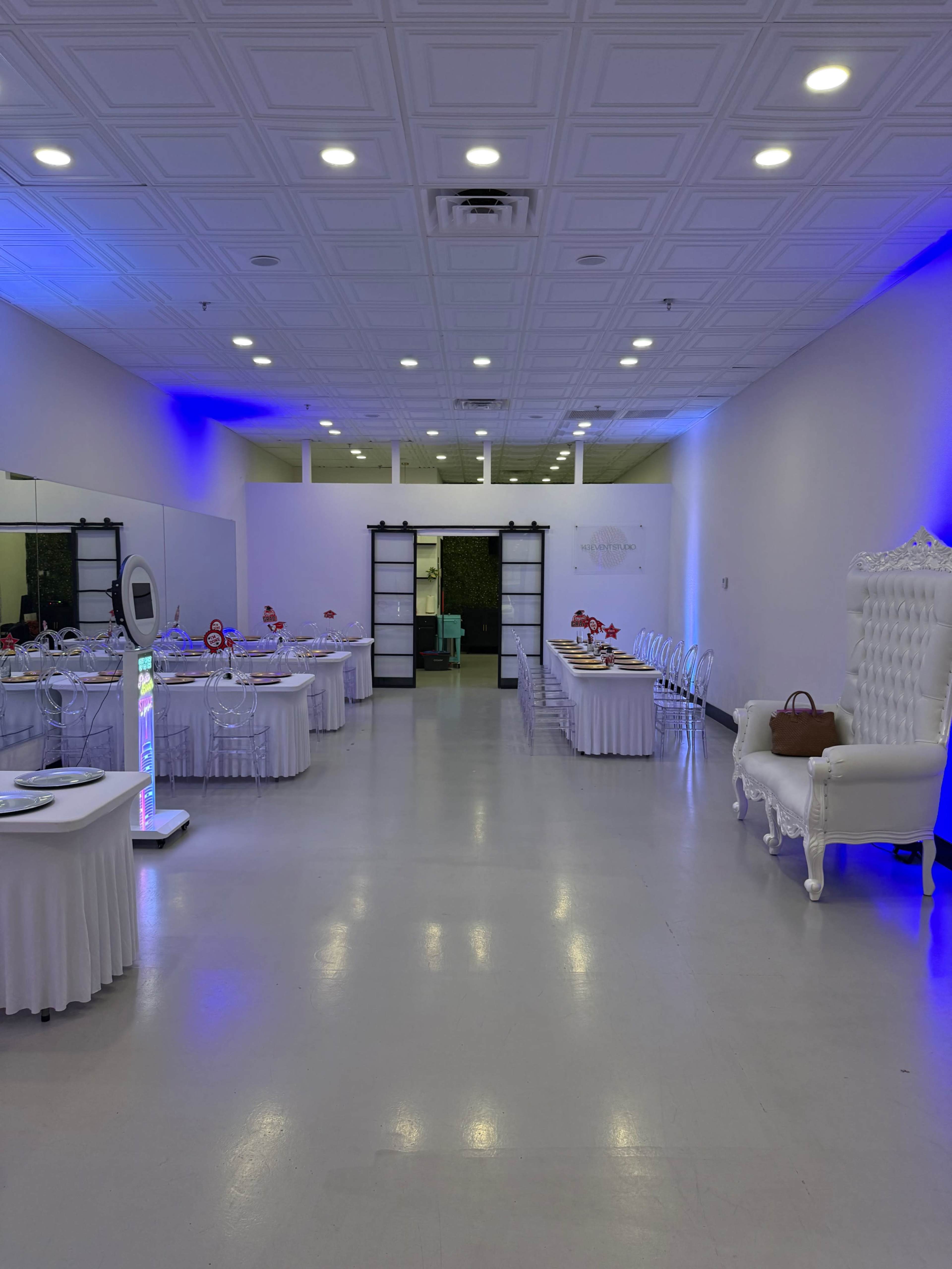 The image shows a spacious event venue with white tables set around the room, illuminated by blue lighting, and featuring a large white throne chair at one end.