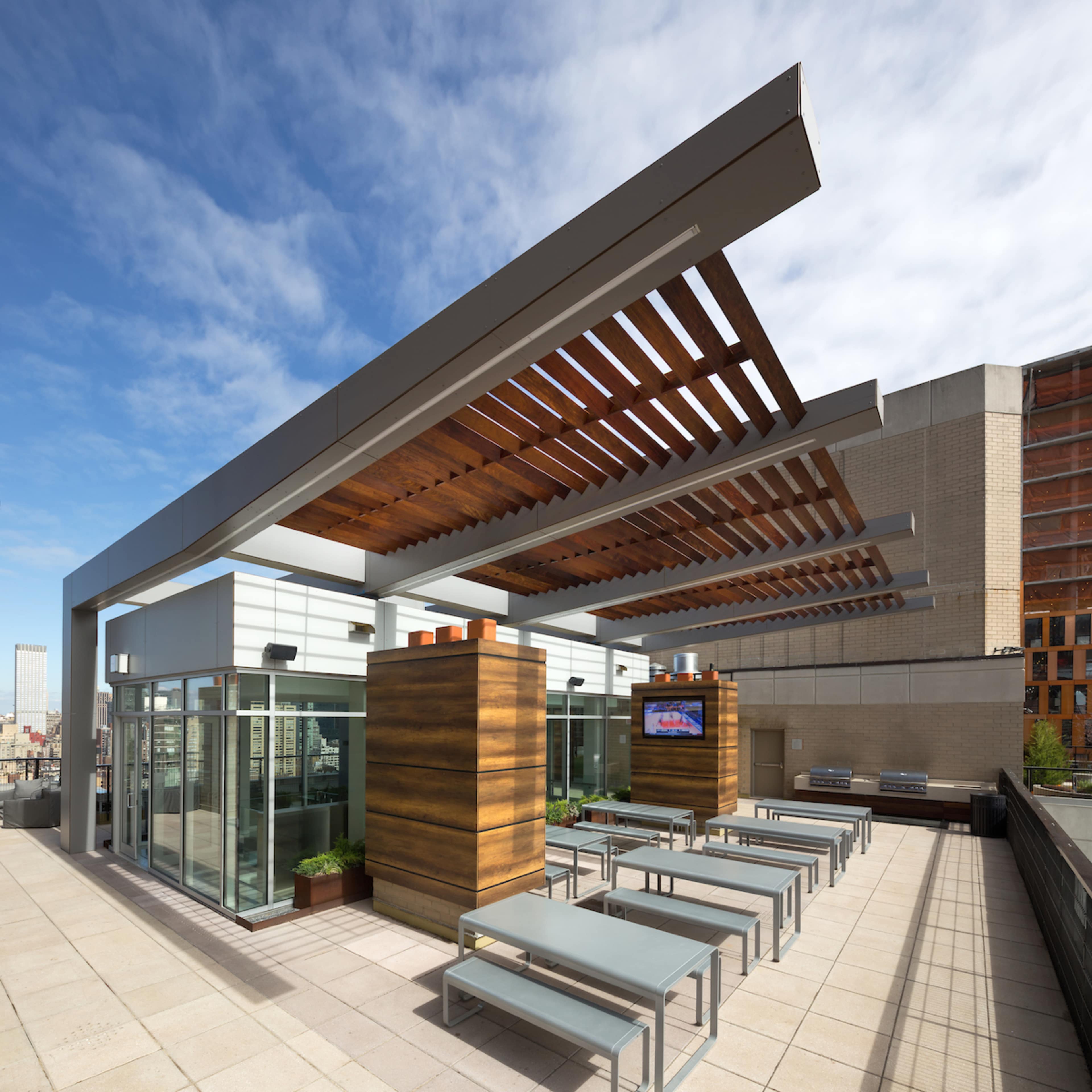 A modern rooftop terrace features a shaded seating area with metal tables and wooden structures under a clear blue sky.