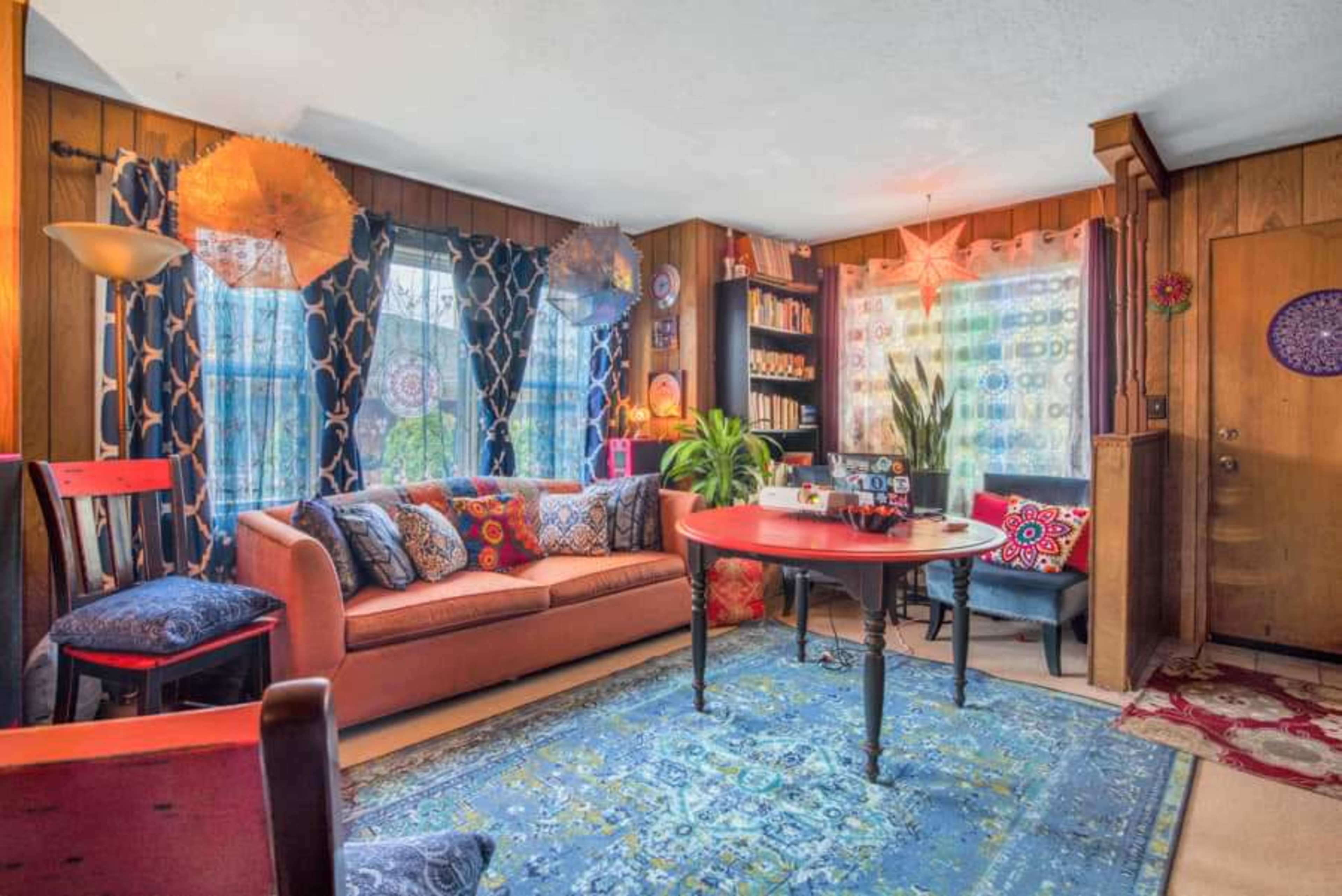 The image shows a colorful living room with a large orange sofa, a round red table, and patterned curtains, surrounded by various decorative items and plants.