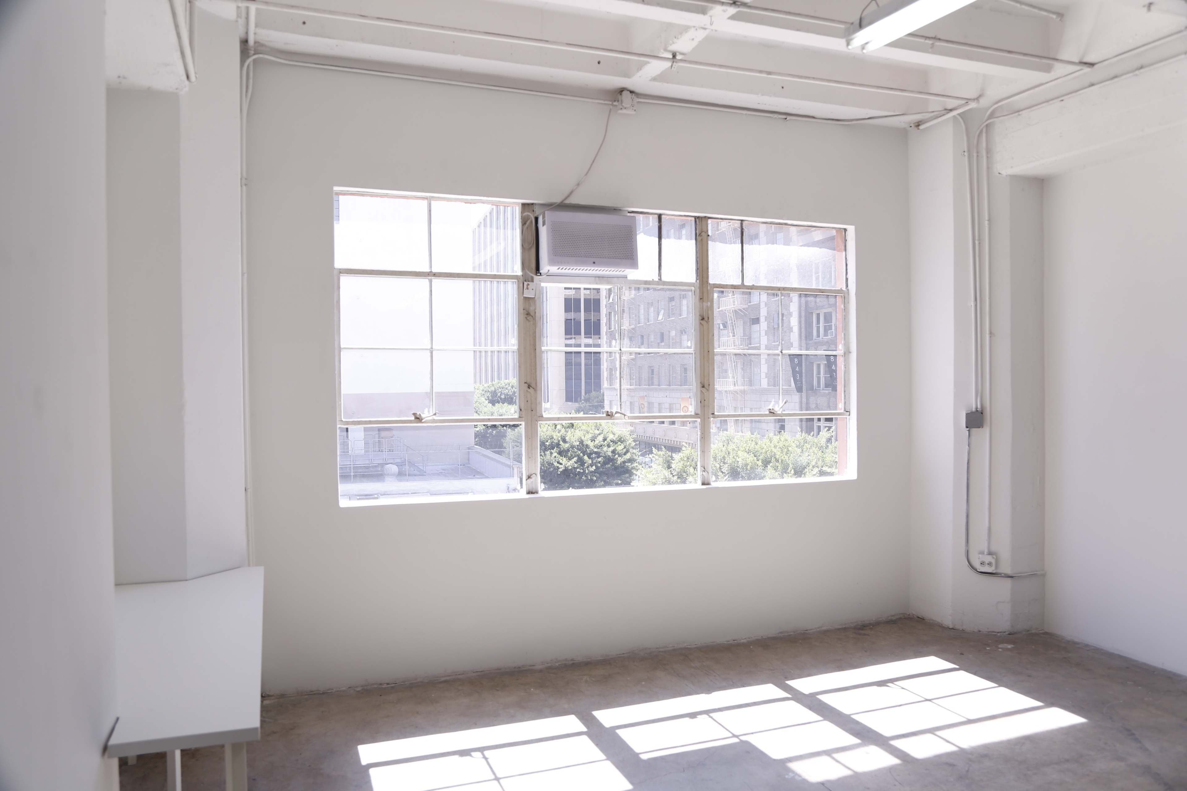 A minimalistic white room with large windows allowing natural light to enter, revealing a cityscape outside.
