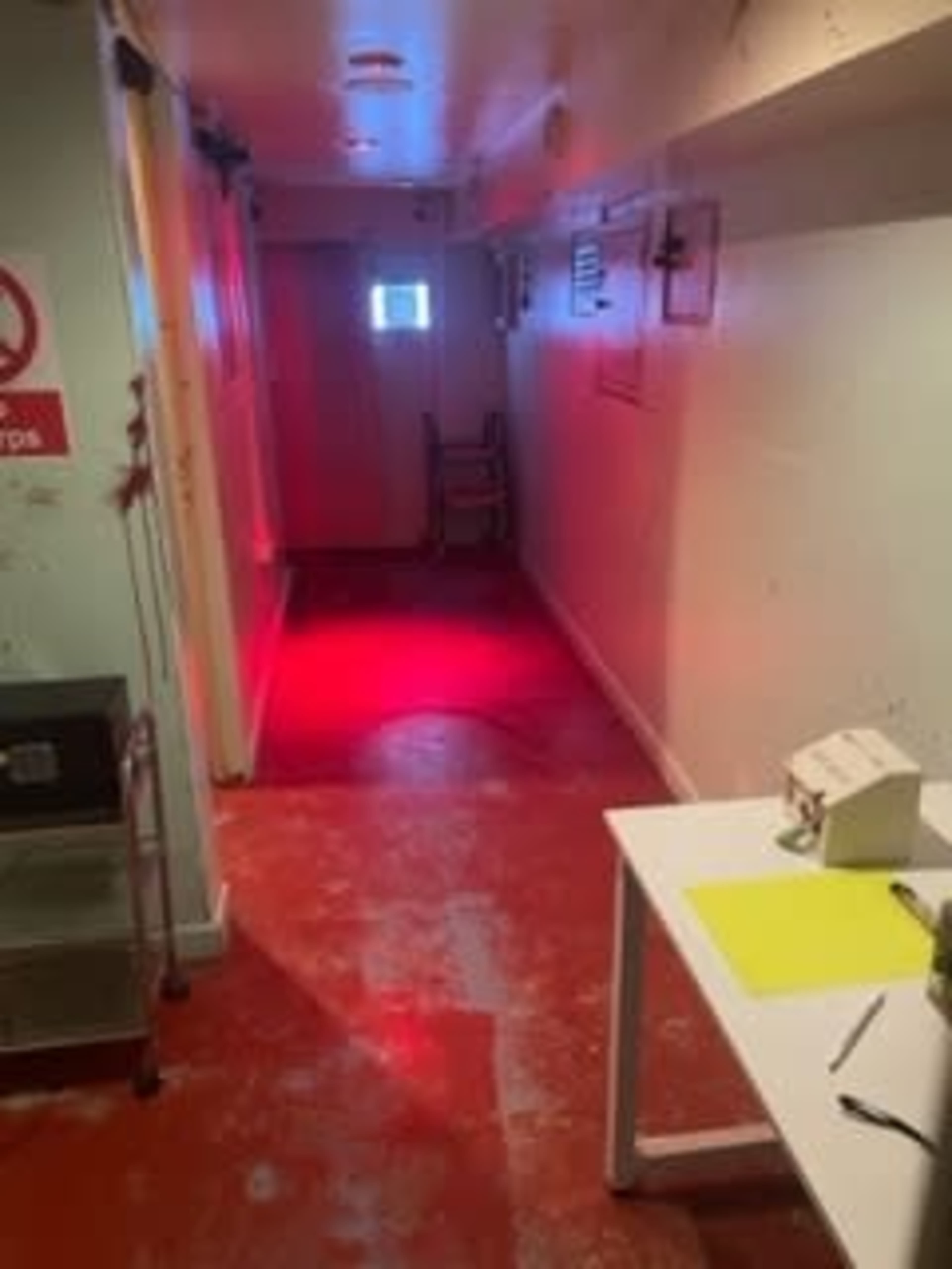 A narrow hallway with red lighting, a window at the far end, and a chair placed against the wall.
