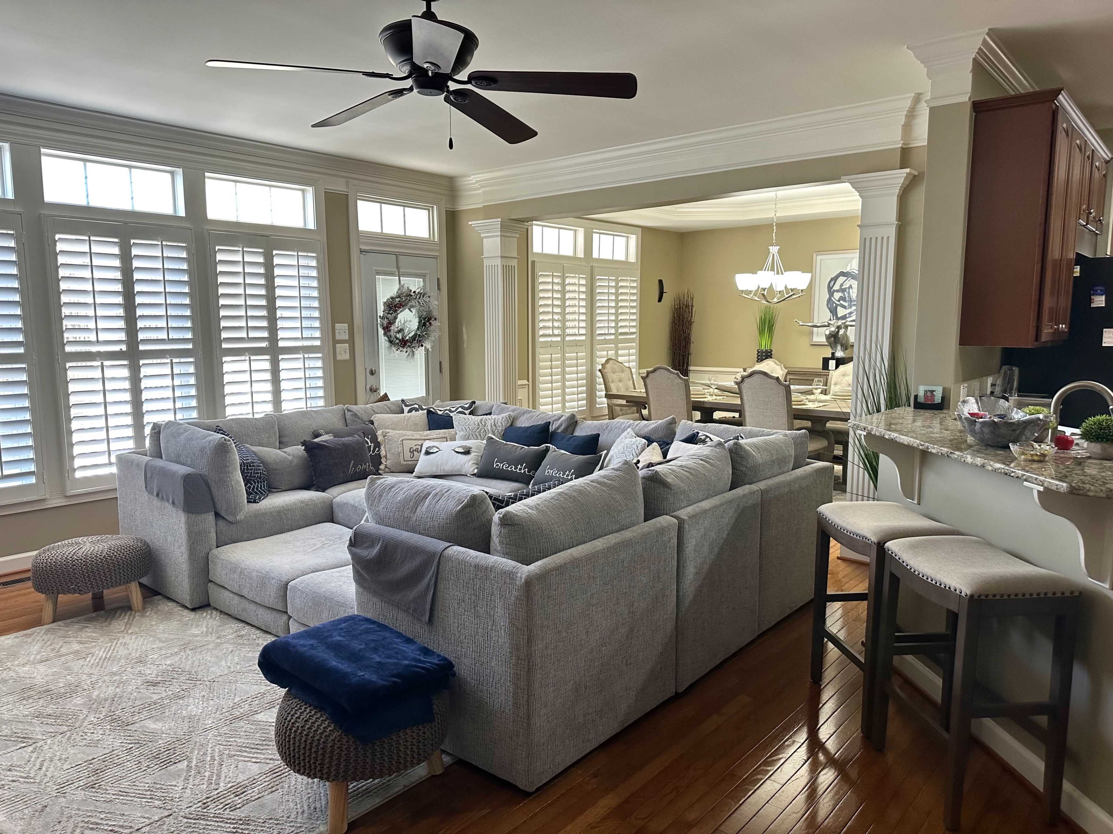 A spacious living room features a large sectional sofa with various cushions, a ceiling fan, and a view into a dining area with a chandelier and a table set for several people.