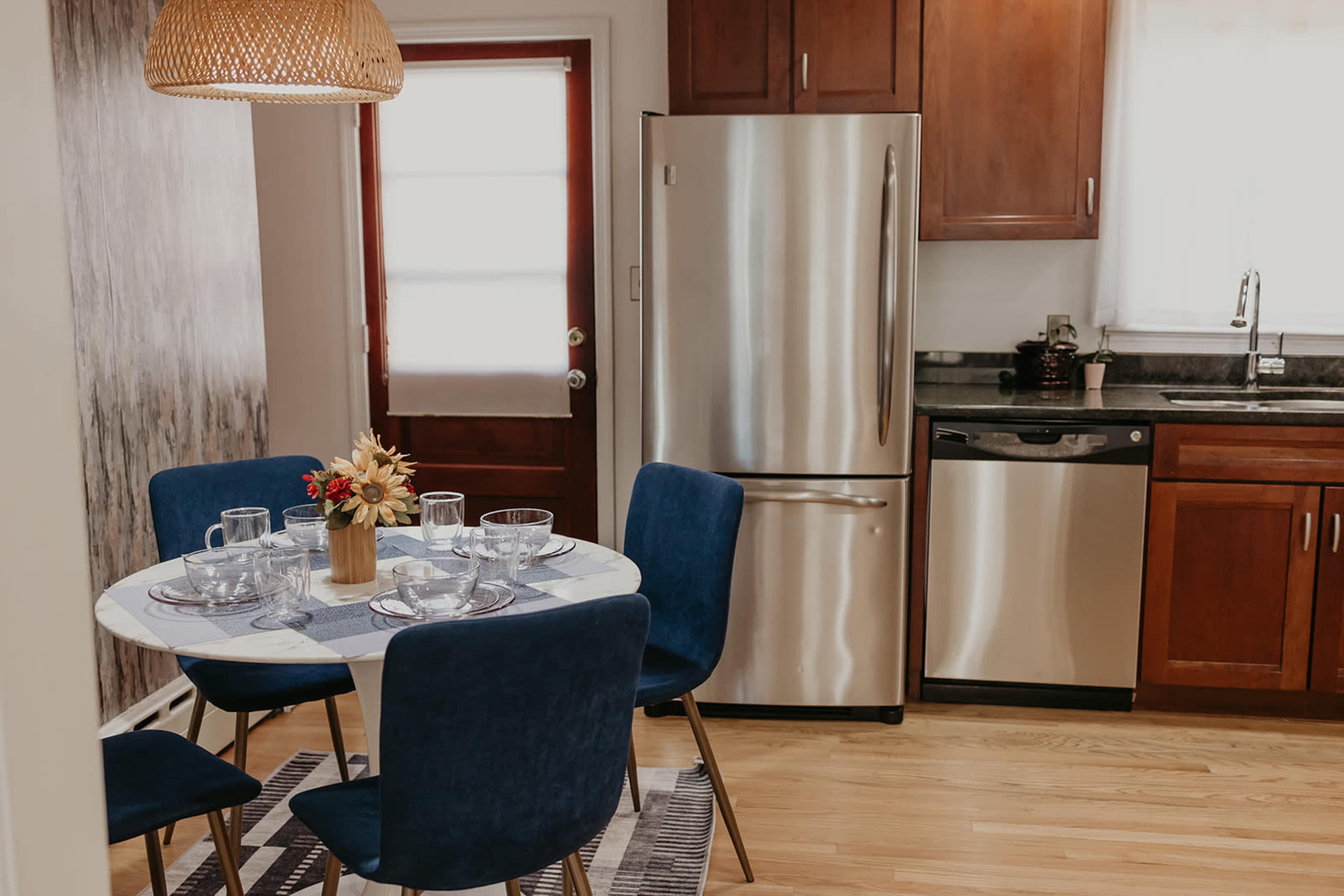 A small dining area features a round table set with glassware and a floral centerpiece, surrounded by blue chairs, beside a modern kitchen with stainless steel appliances.