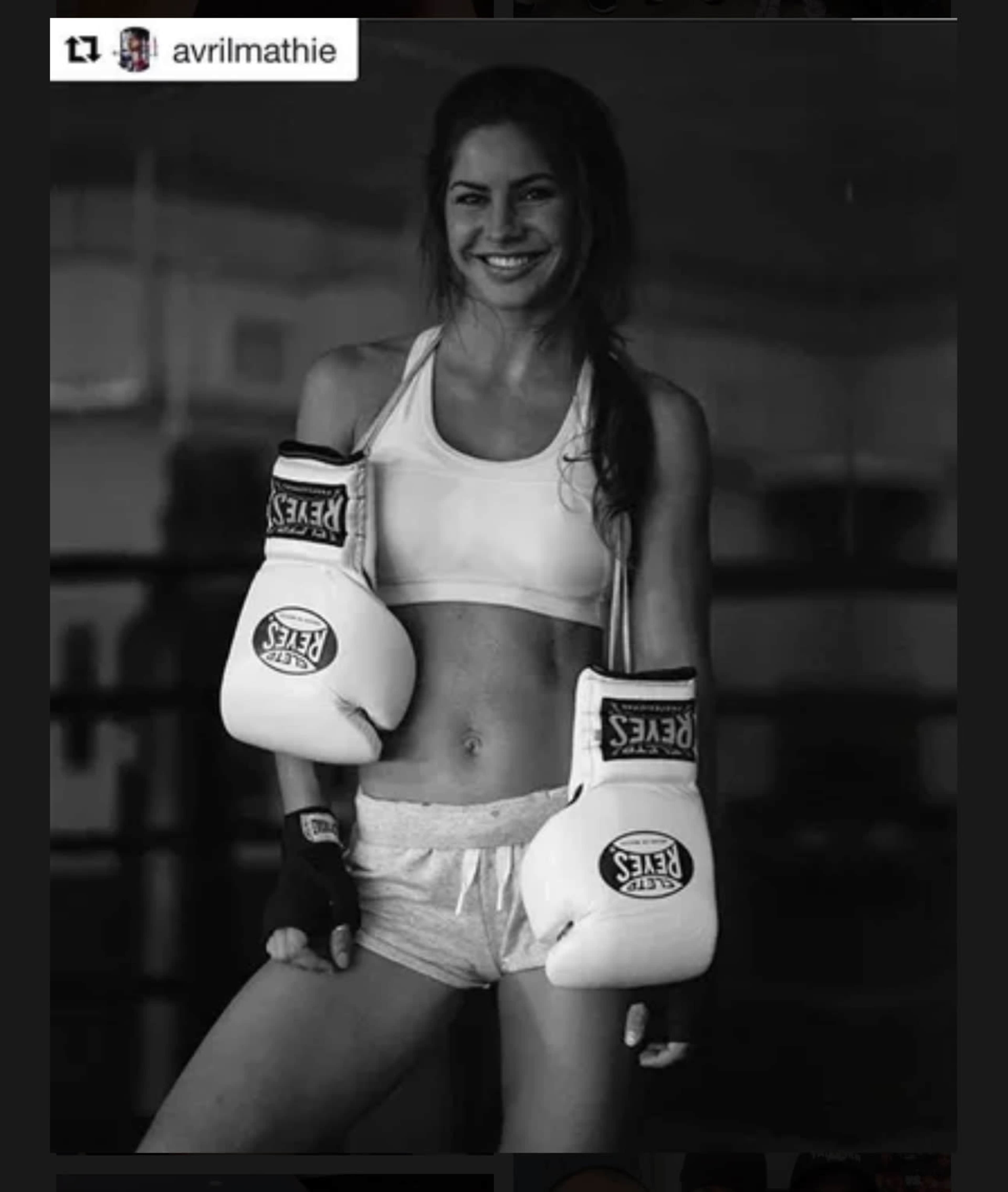 A woman poses in a boxing gym wearing white boxing gloves and sporting athletic wear.