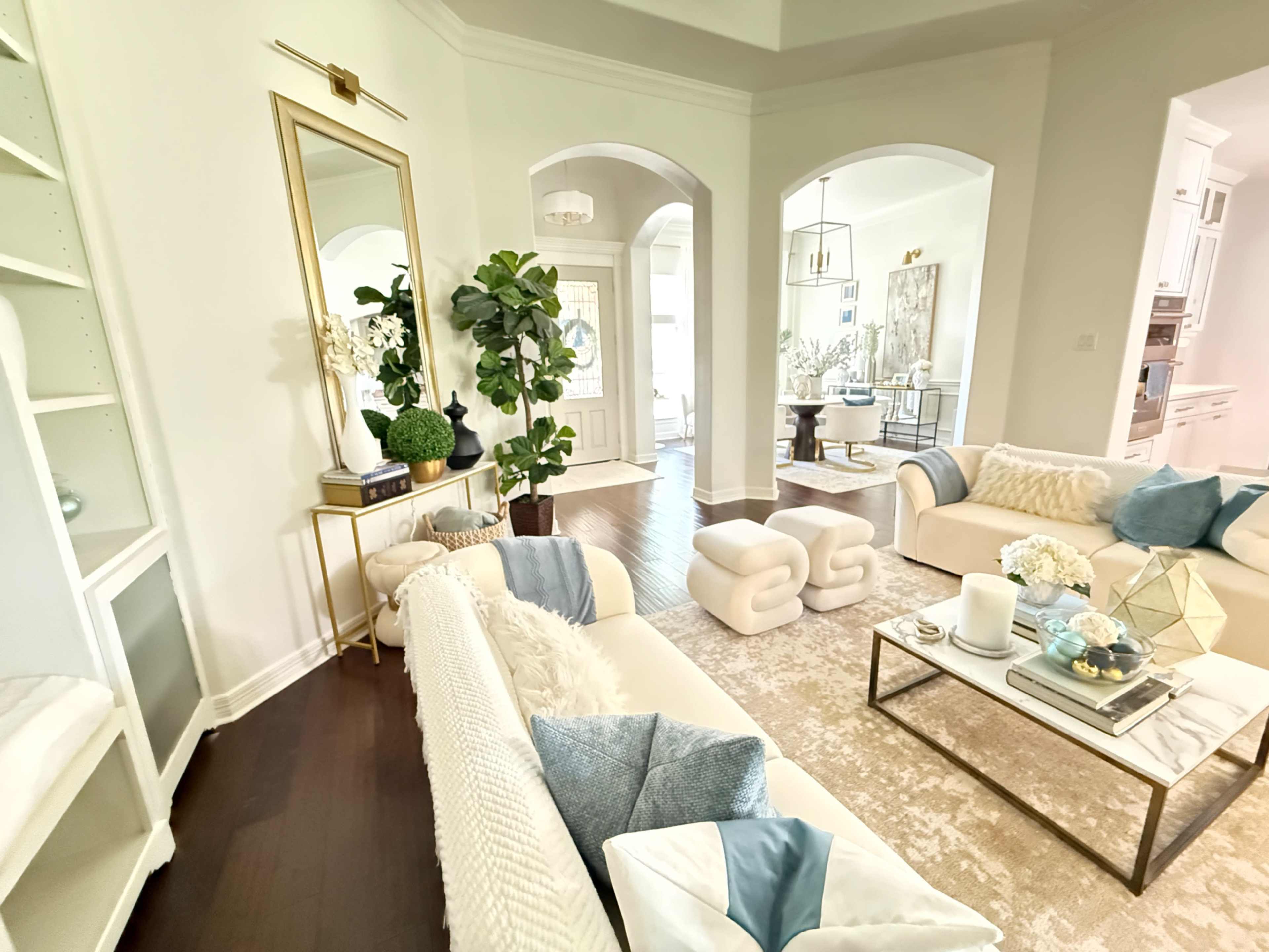 The image shows a modern living room with light-colored sofas, a coffee table, and a large mirror, leading to an open dining area.