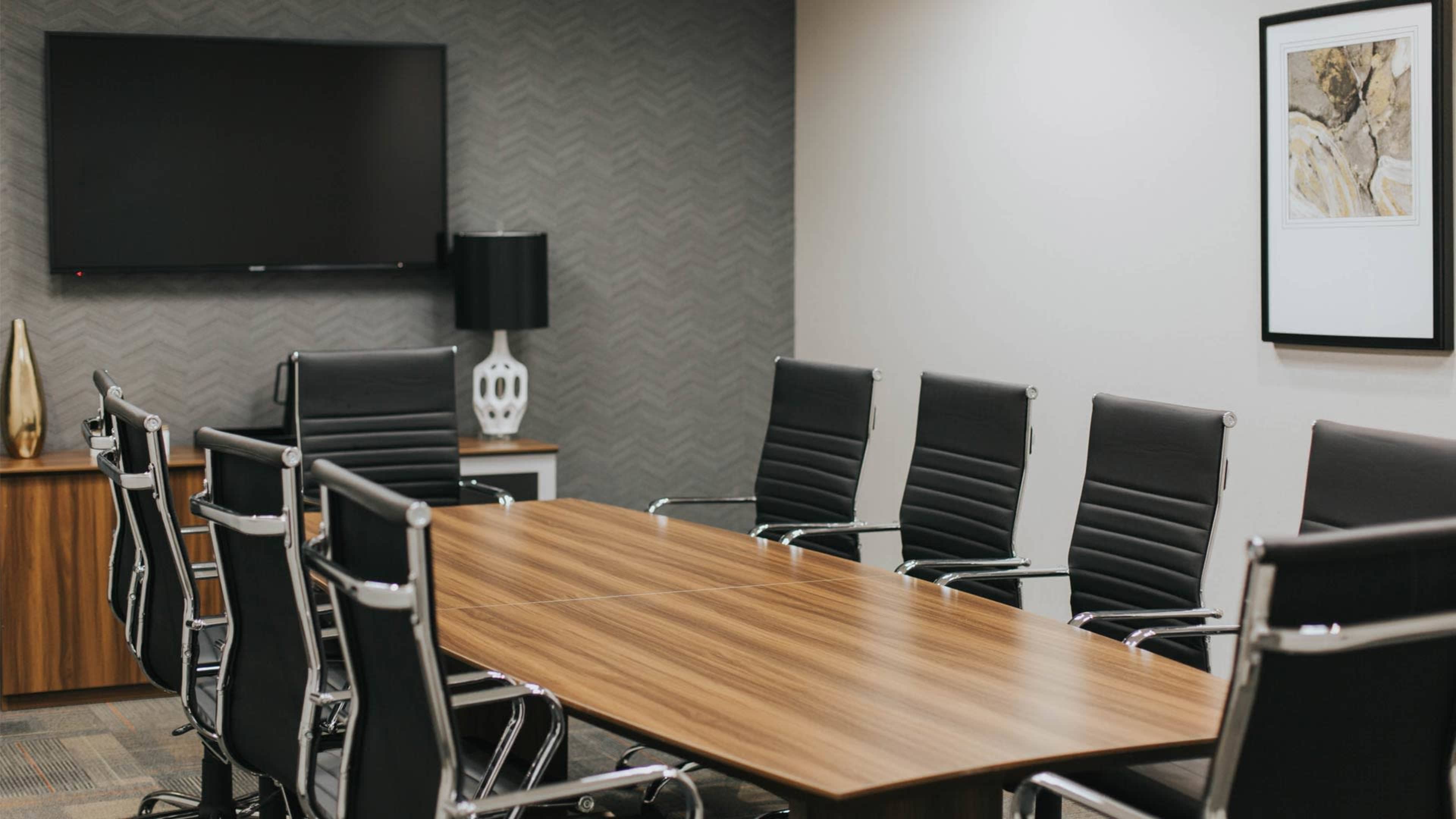 A modern conference room features a wooden table surrounded by black leather chairs, with a television and framed artwork on the walls.
