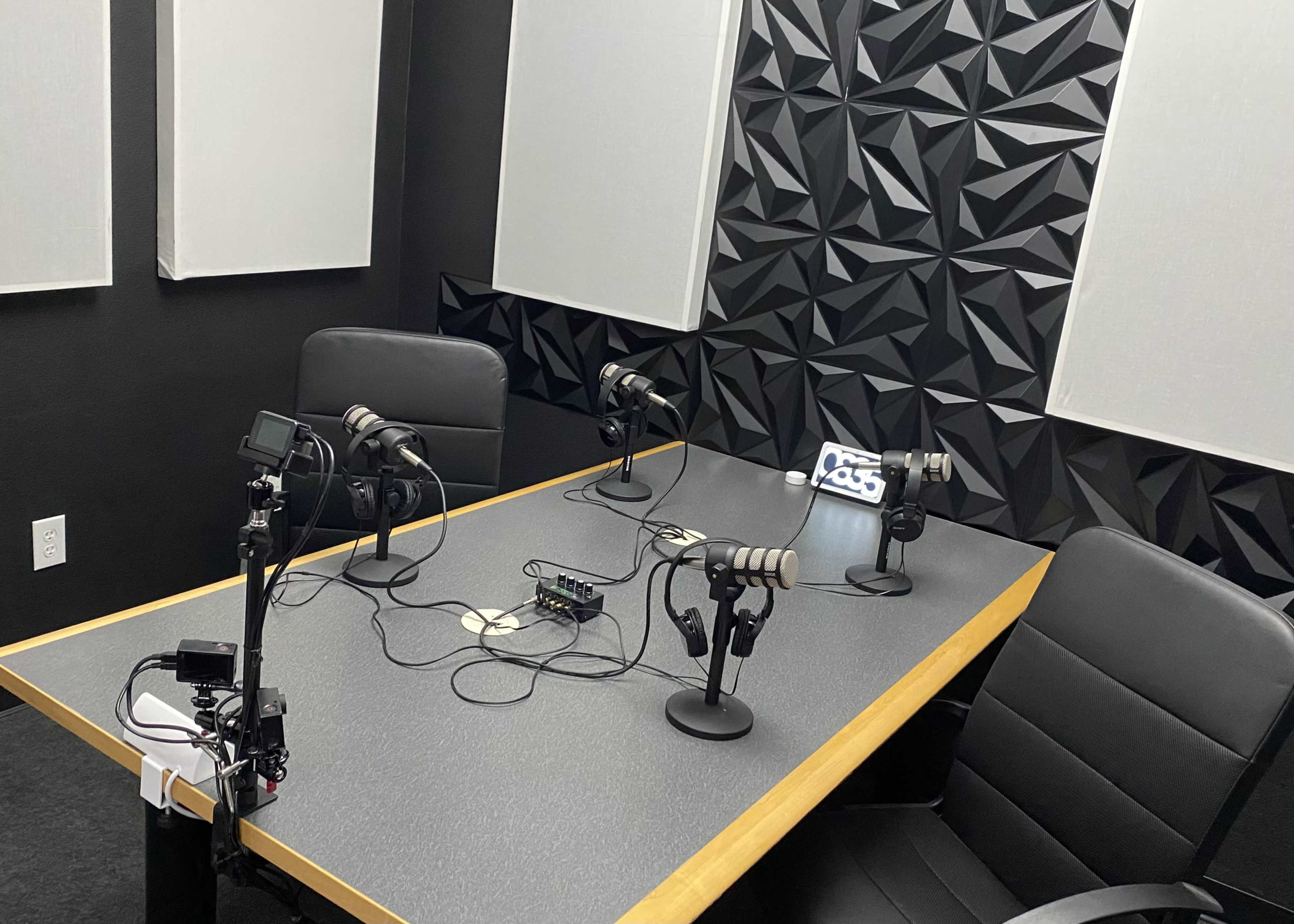 The image shows a recording studio with multiple microphones arranged around a table, set against a textured black wall and equipped with audio devices.