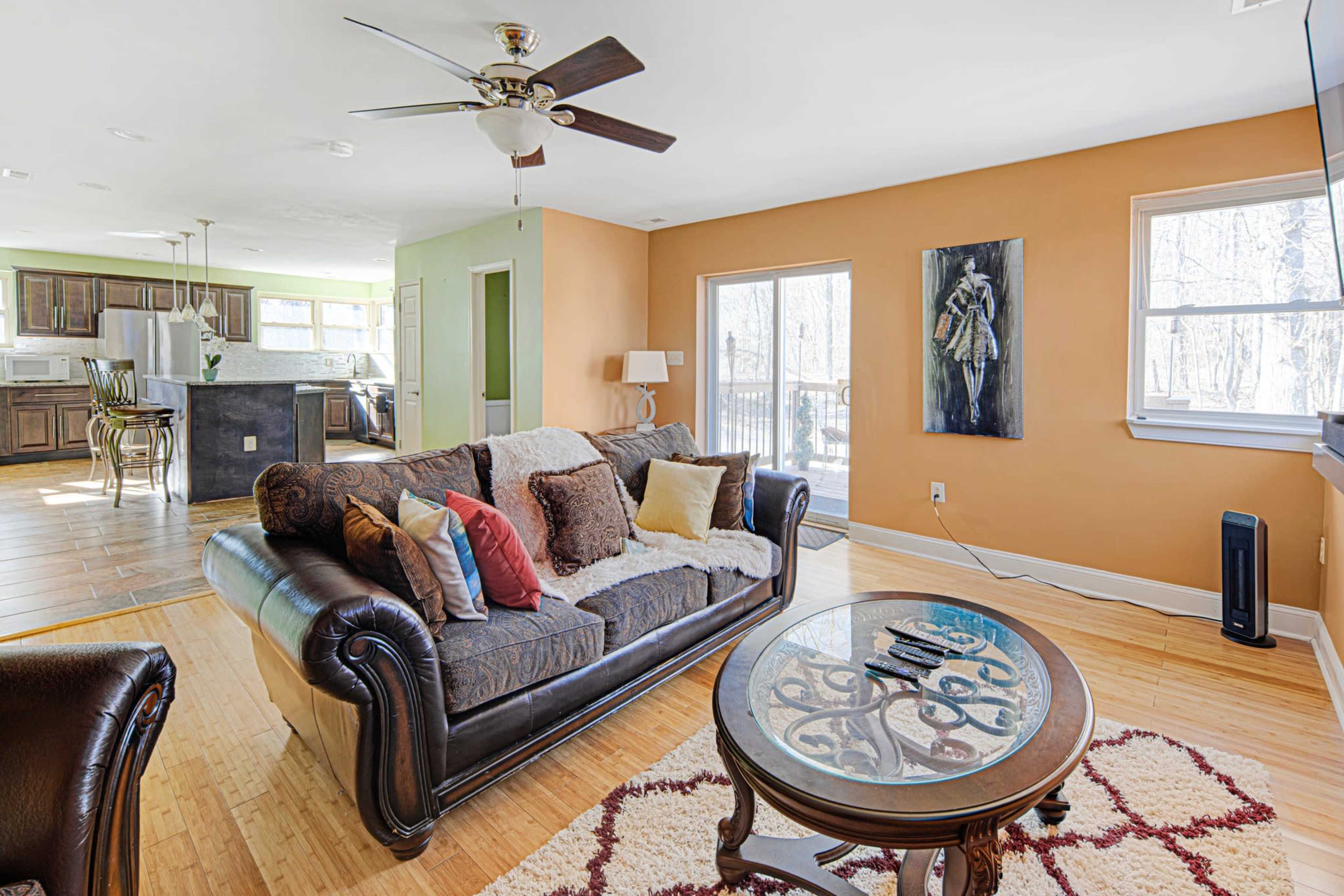 A cozy living room features a dark leather sofa with decorative pillows, a round coffee table, and large windows that provide natural light, with an open kitchen visible in the background.