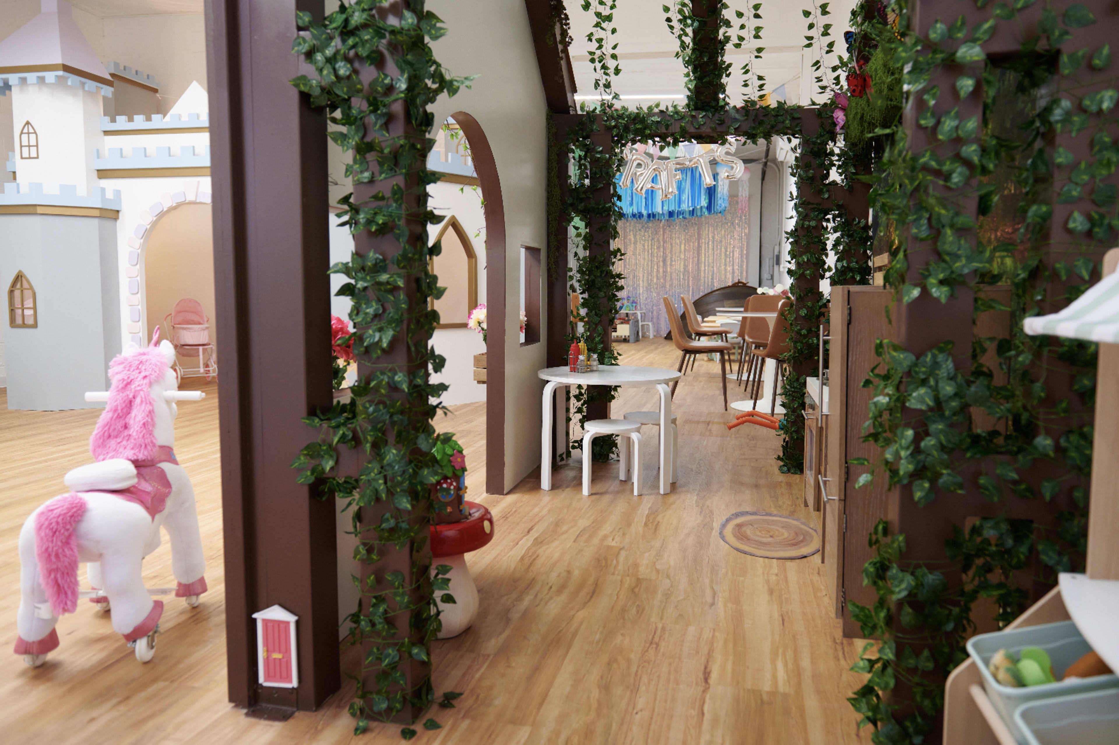 The image shows a whimsical indoor play area featuring castle-like structures, greenery on the walls, and a seating arrangement with a table and piano.