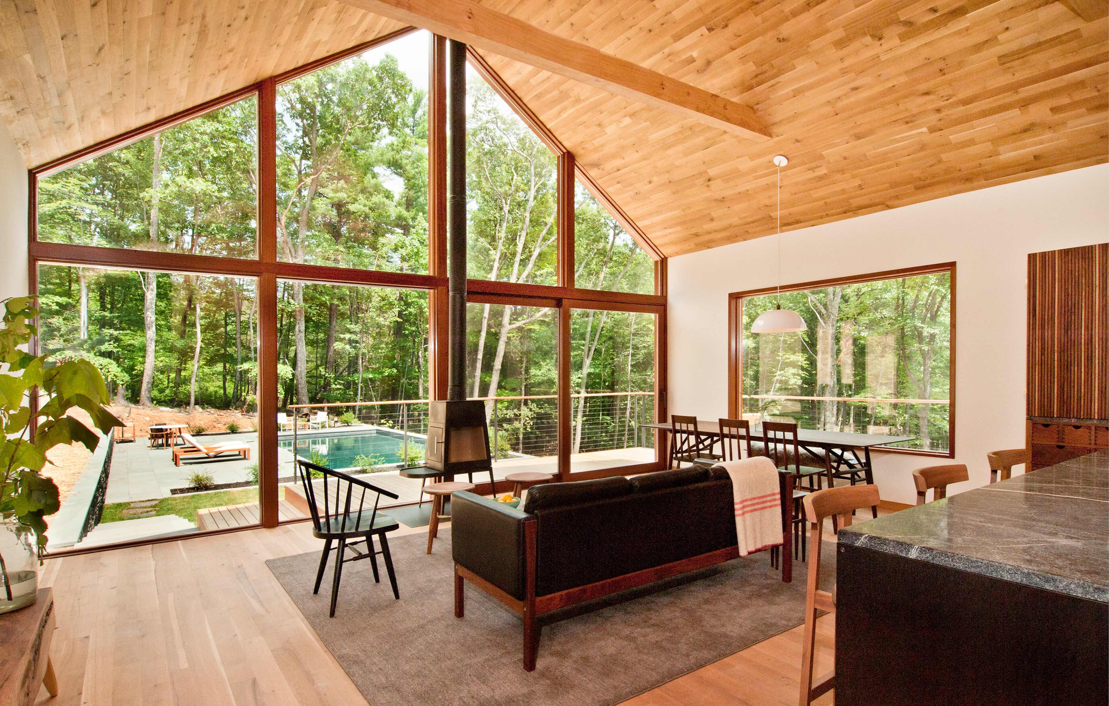 A modern living area features large windows overlooking a pool and wooded landscape, with a dining table and stylish furniture.