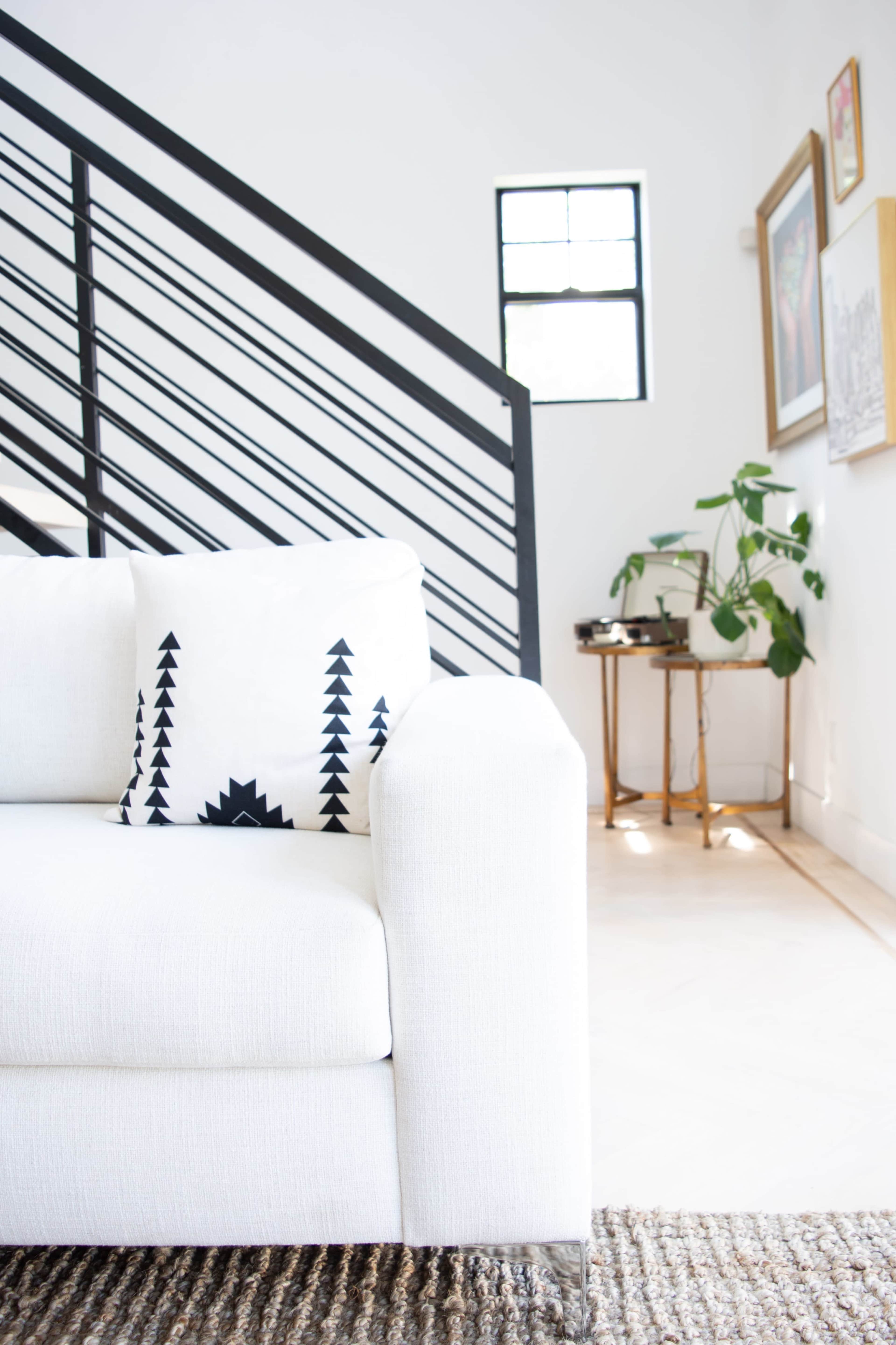 A white sofa with a patterned pillow is positioned next to a staircase and a small side table with a plant.