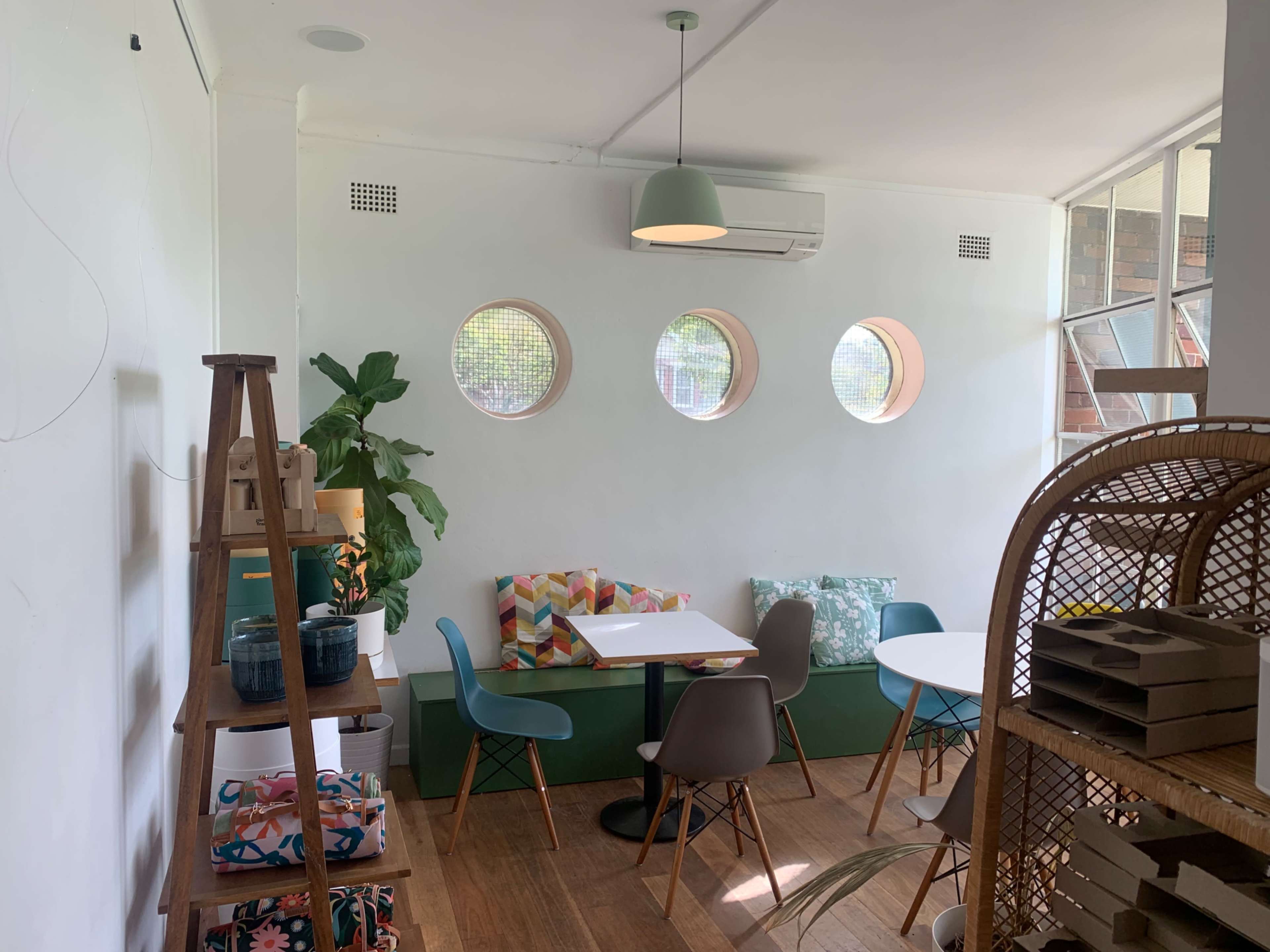 The image shows a bright cafe interior with circular windows, a small table with chairs, and a decorative shelf featuring plants and colorful cushions.