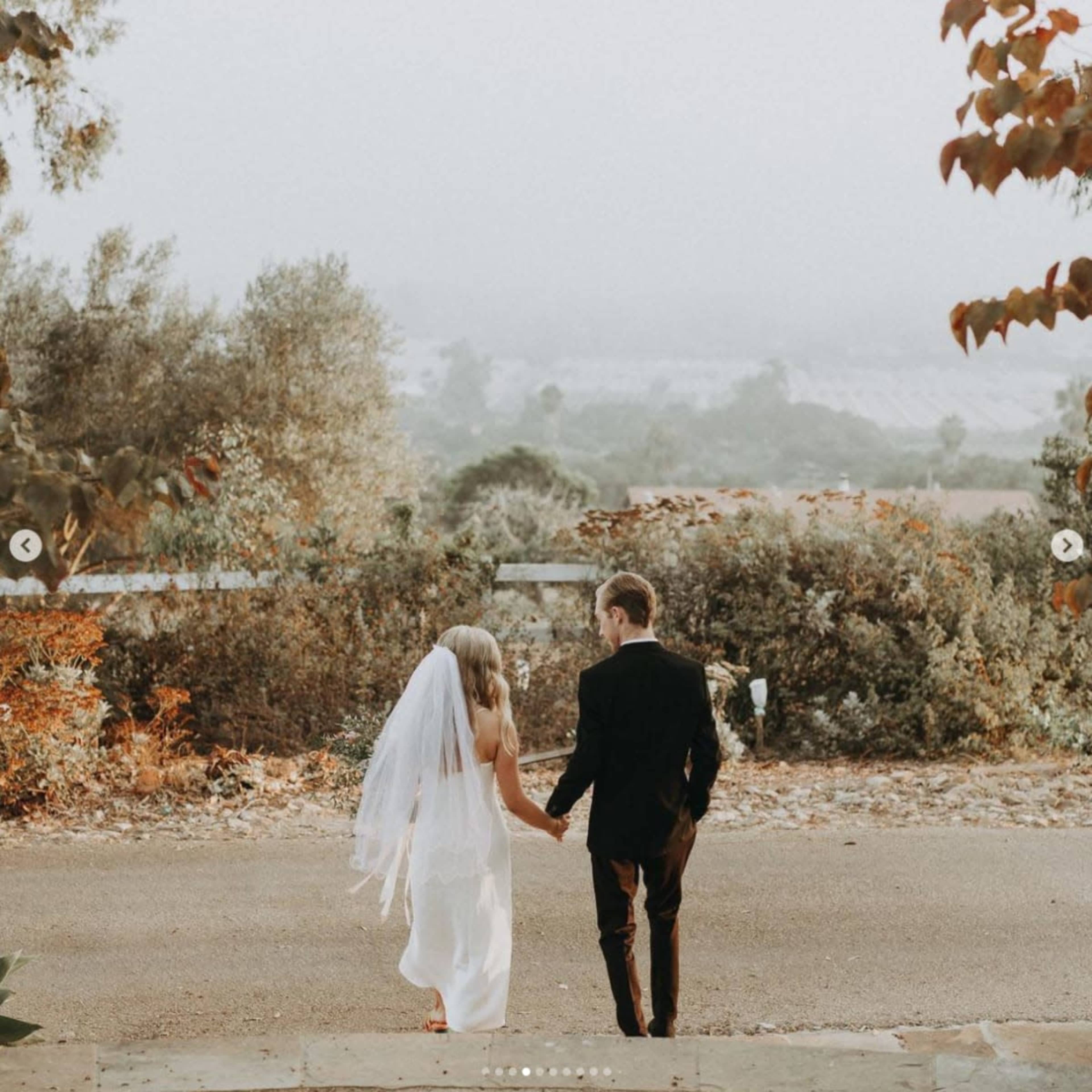 A bride in a white dress and a groom in a black suit walk hand-in-hand down a path with a scenic background of trees and hills.
