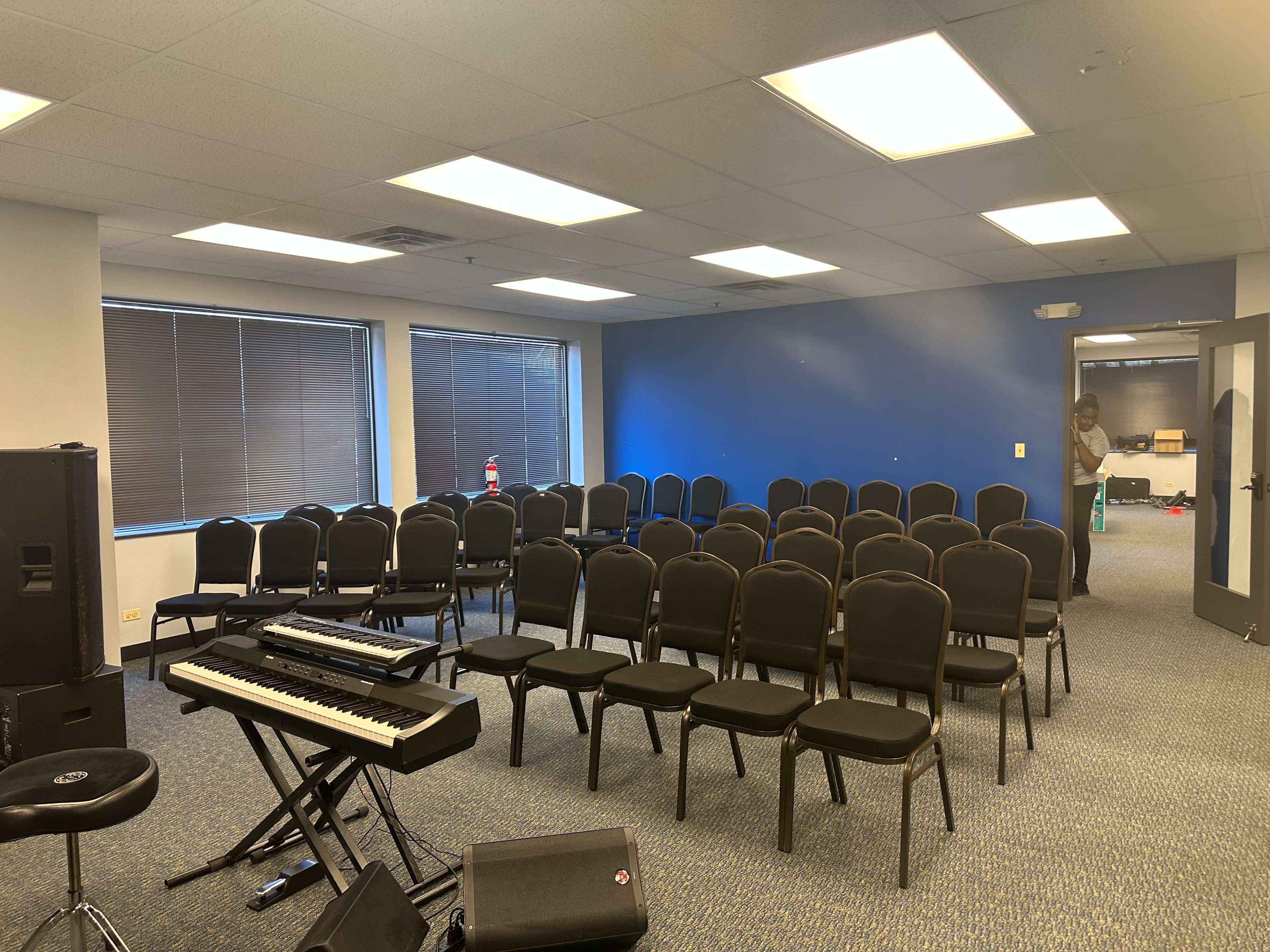 The image shows a small room set up with rows of black chairs facing a keyboard, with a person standing in the background.