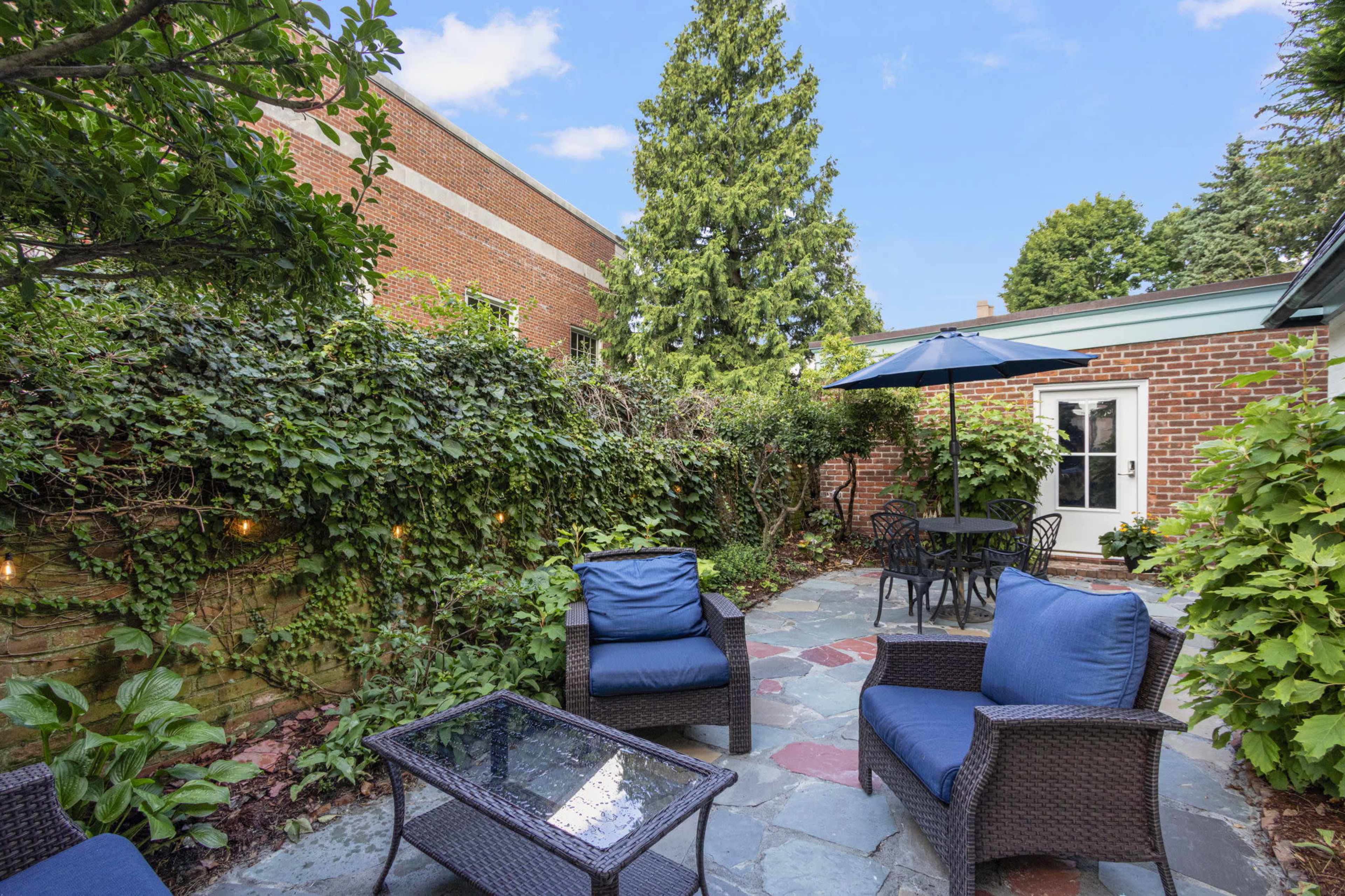 A cozy outdoor seating area features blue chairs and a table surrounded by greenery and a small patio area, with a large umbrella providing shade.