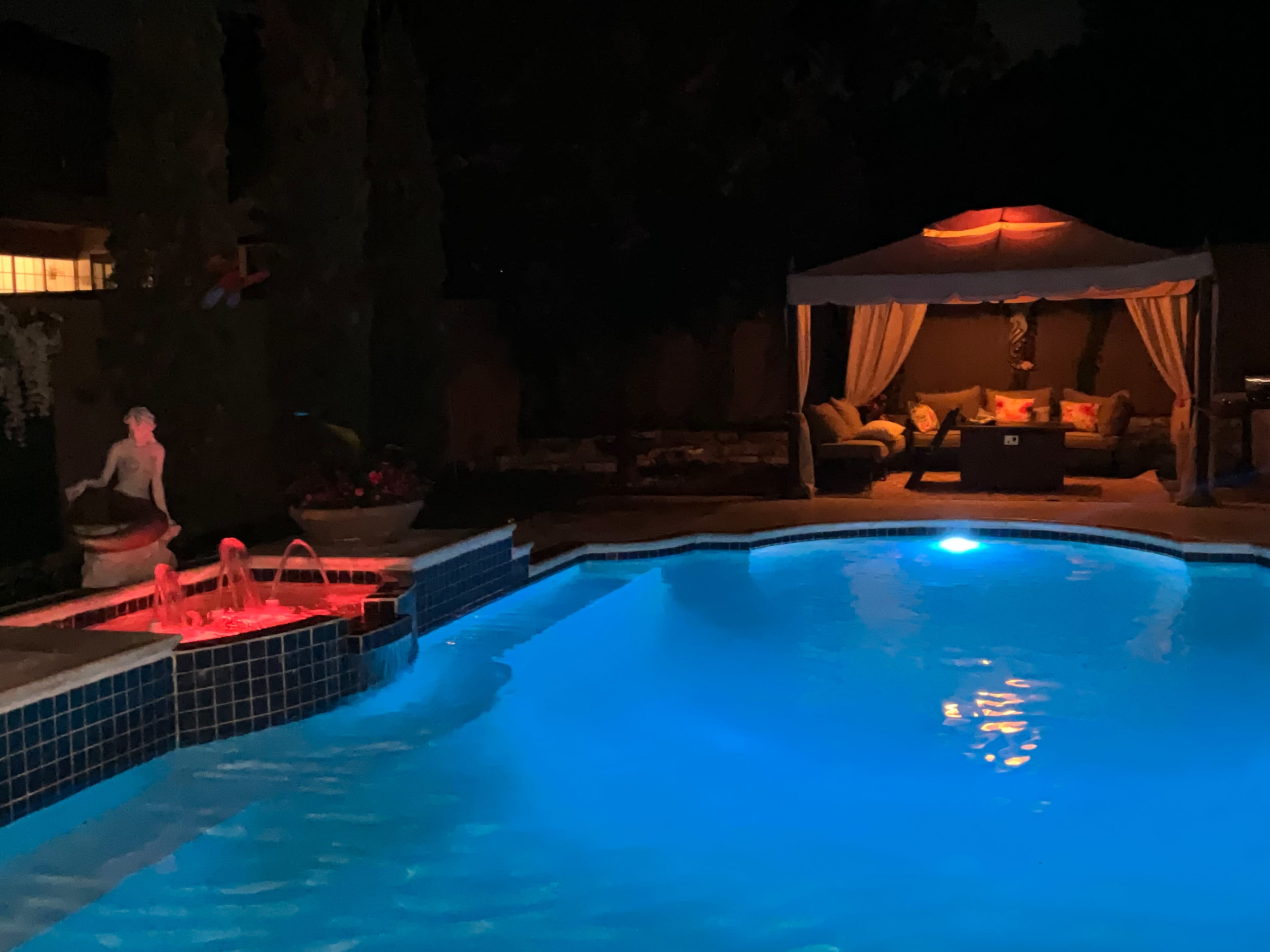 A well-lit pool at night with a water feature and a tented seating area beside it.
