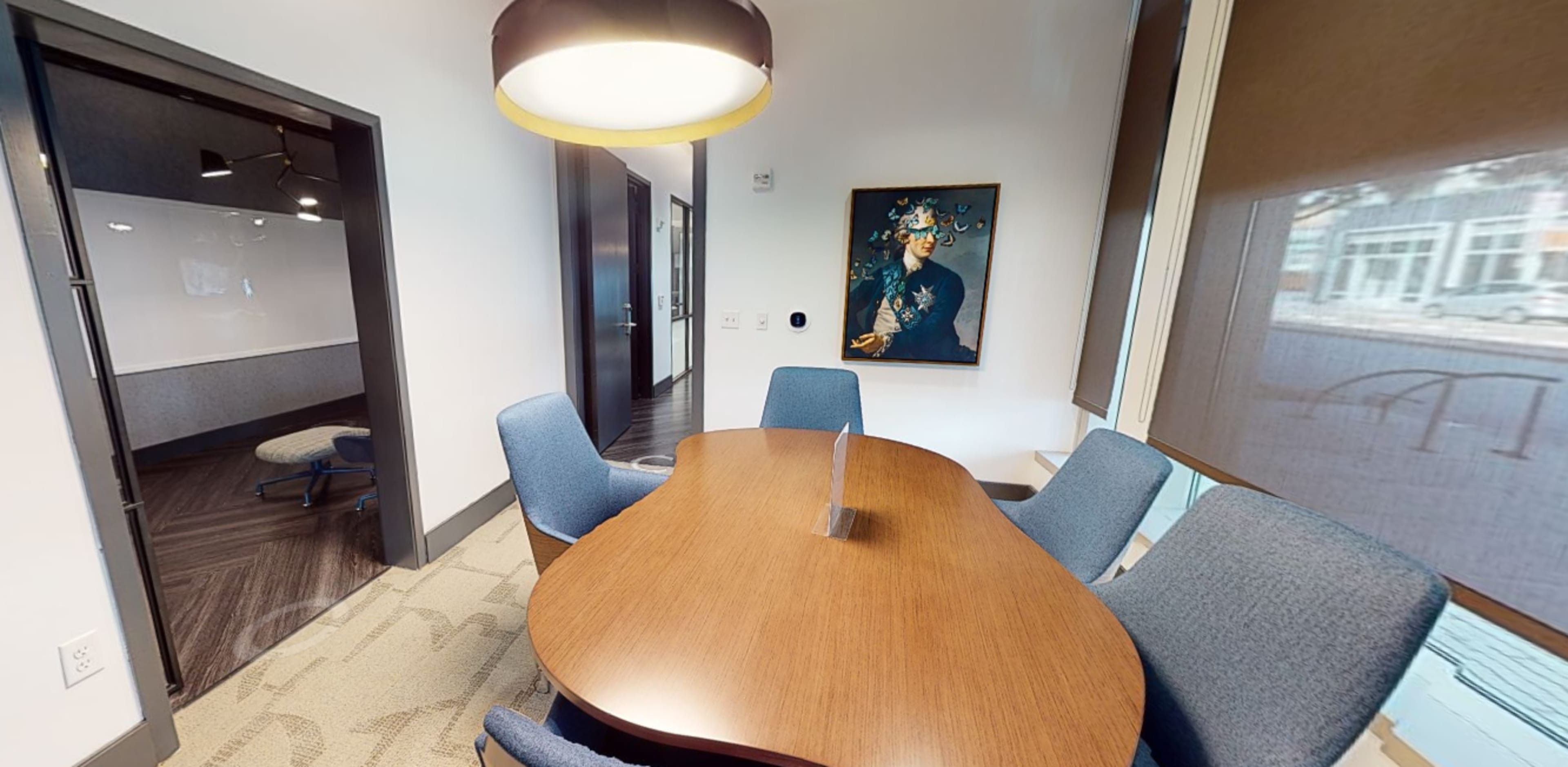 A modern conference room features a round wooden table surrounded by blue chairs, with a framed painting on the wall and a view of the street outside through large windows.