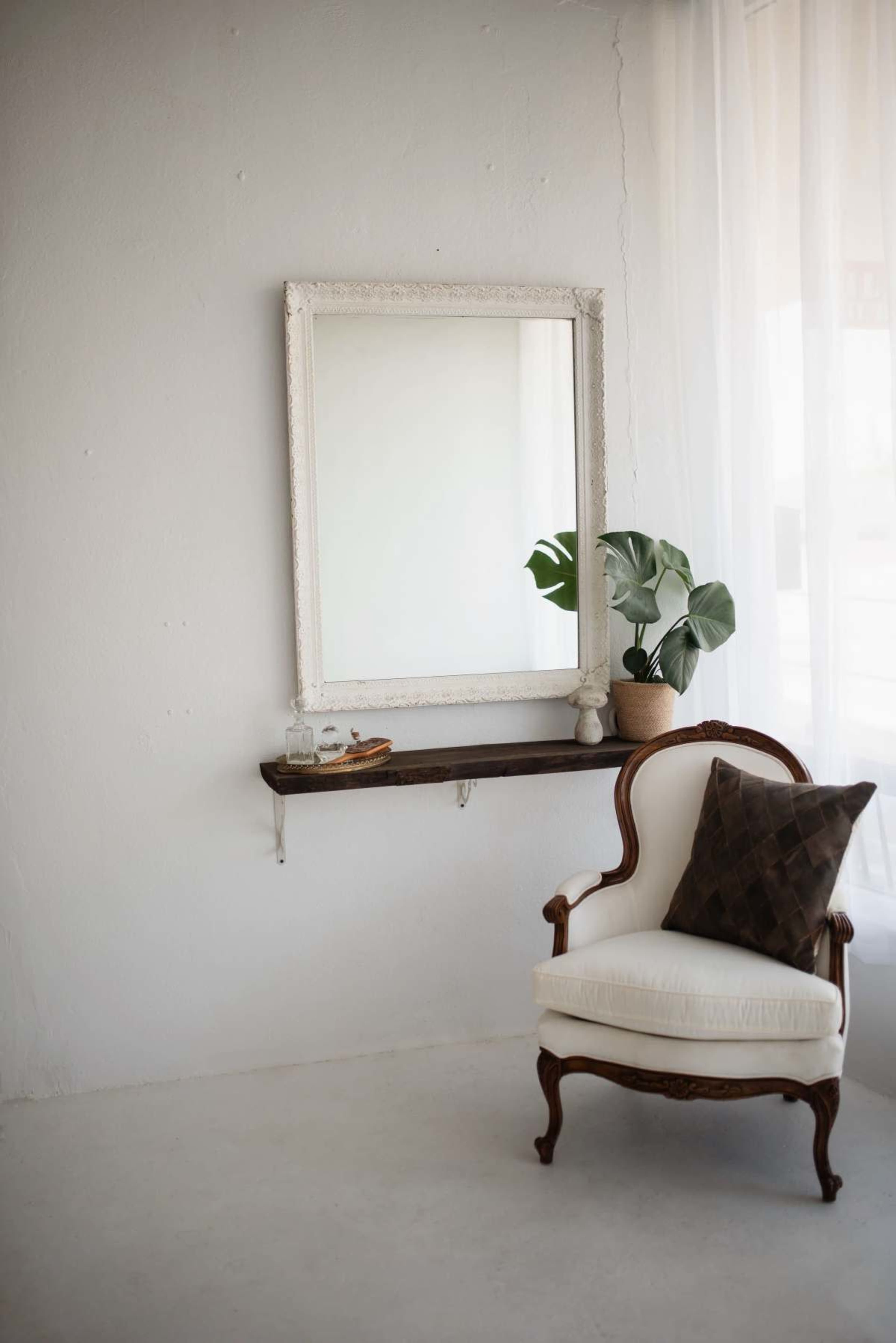 A white wall features a large ornate mirror above a wooden shelf, next to a vintage chair with a decorative pillow and a potted plant.