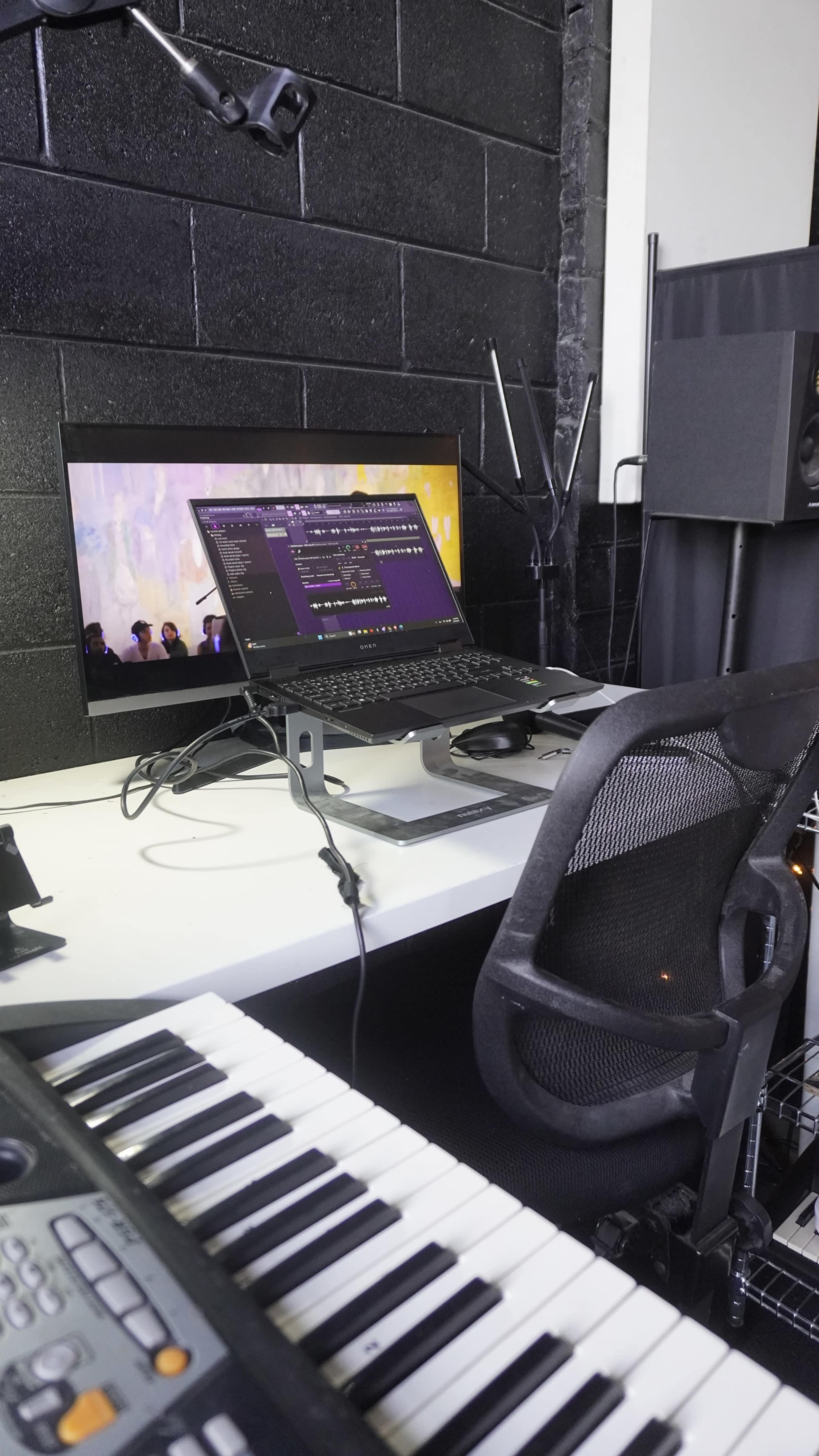 A workspace features a computer monitor displaying music software, a laptop on a stand, a keyboard, and audio equipment against a black wall.