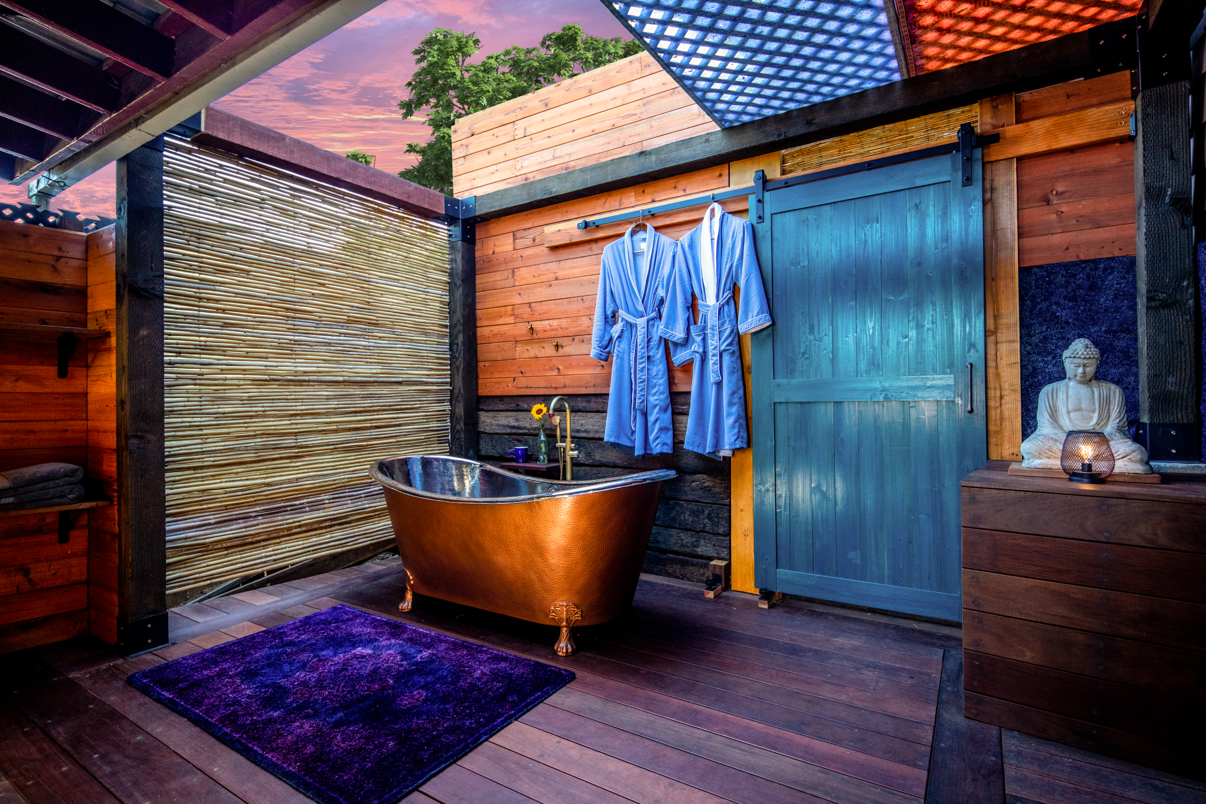 The image shows a wooden outdoor bathroom featuring a copper bathtub, two blue robes hanging on a wall, and a statue of Buddha on a side table.