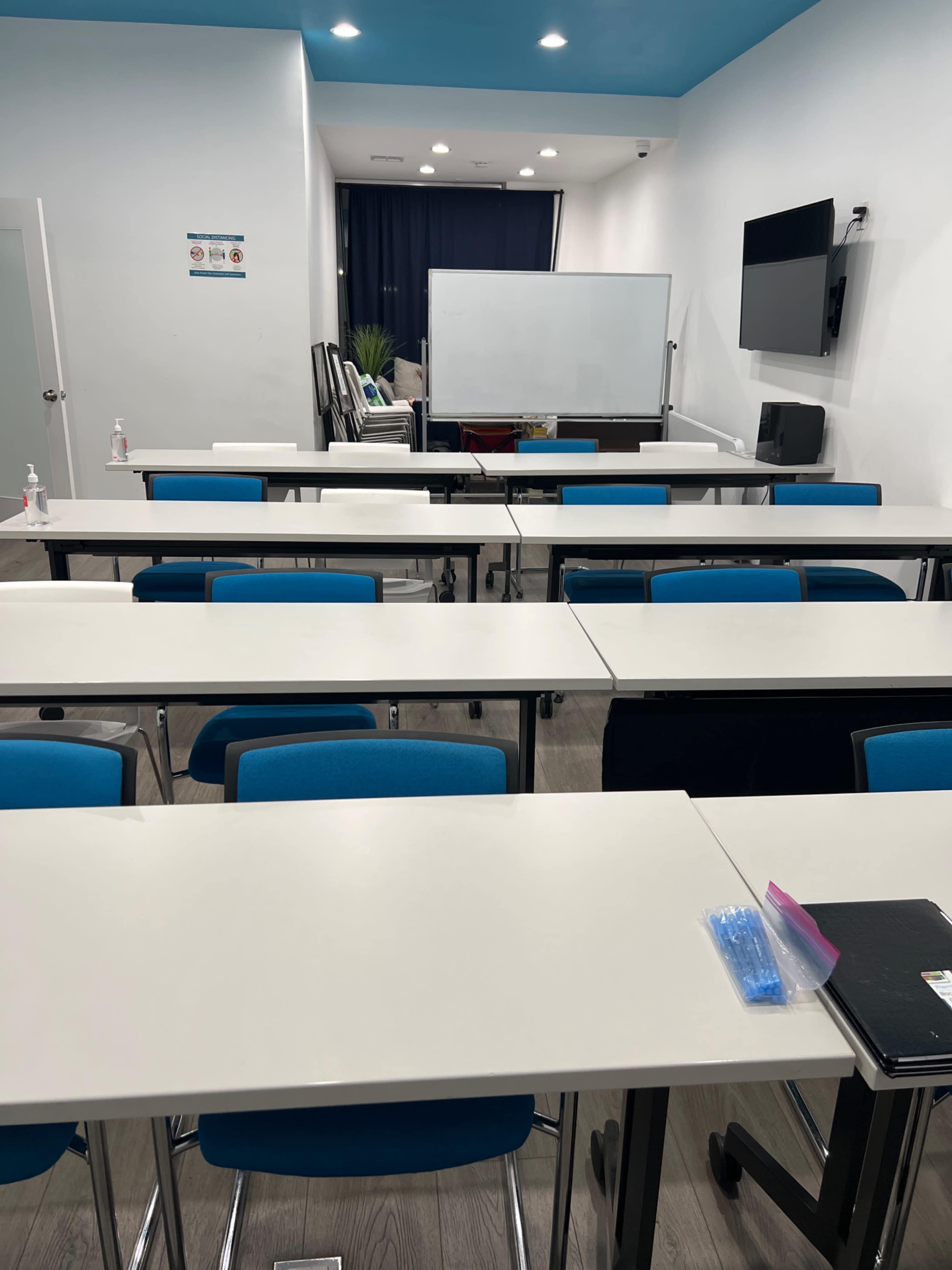 An empty classroom with rows of tables and chairs, a whiteboard at the front, and a television mounted on the wall.