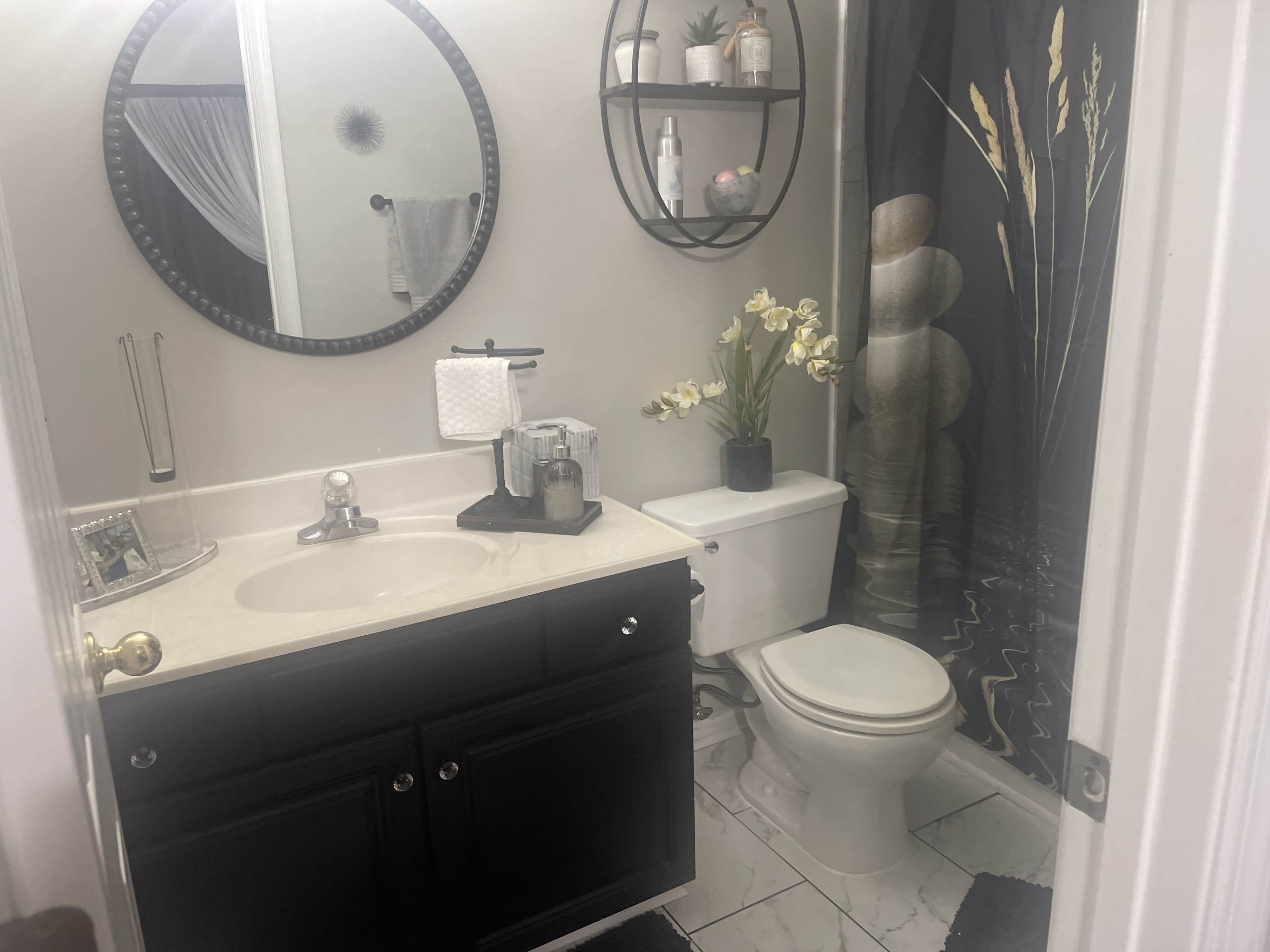The image shows a small bathroom featuring a circular mirror, a sink with dark cabinetry, a toilet, and a shower curtain with a nature-themed design.