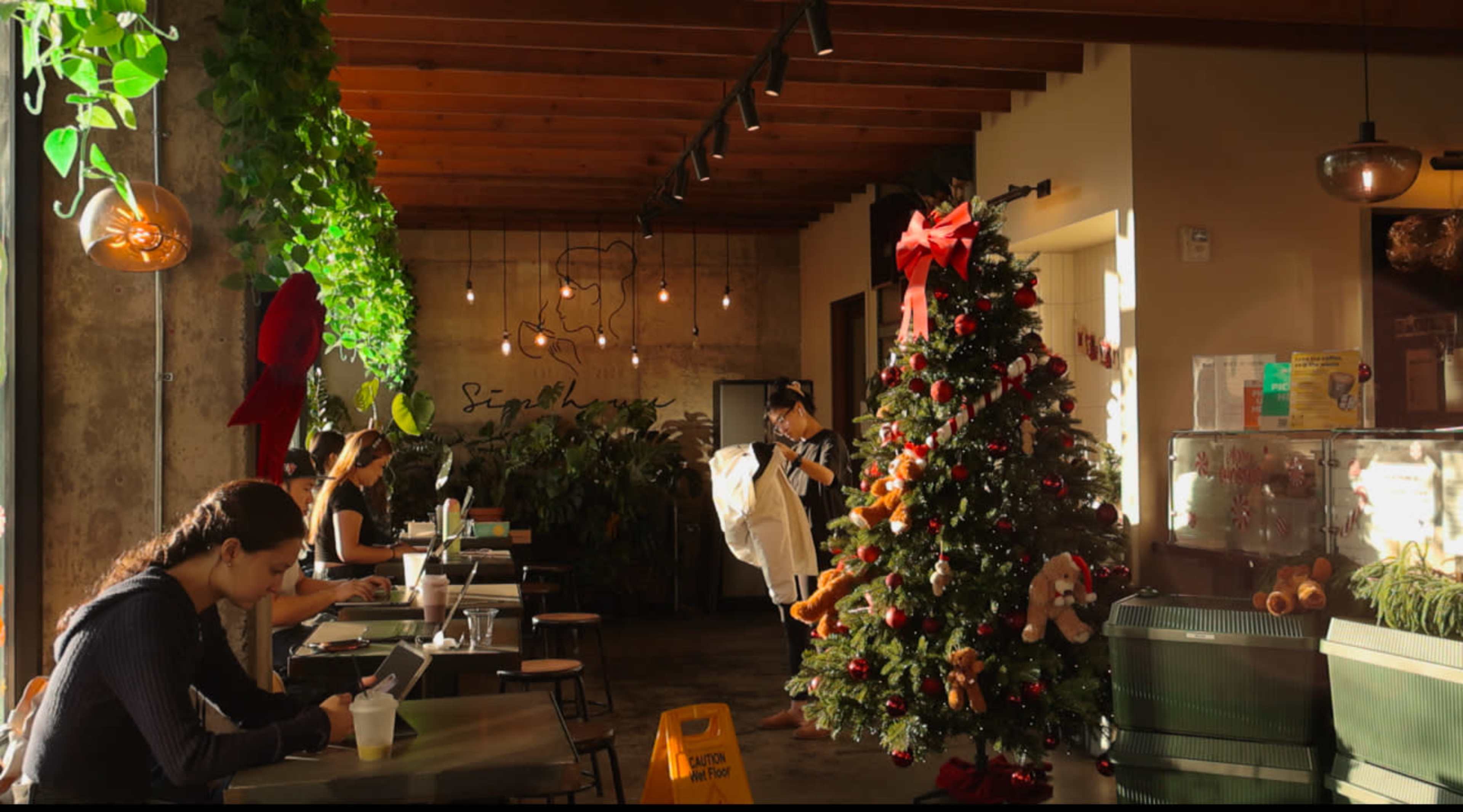 The image shows a café interior adorned with greenery, featuring a Christmas tree decorated with ornaments and a couple of people seated at tables.