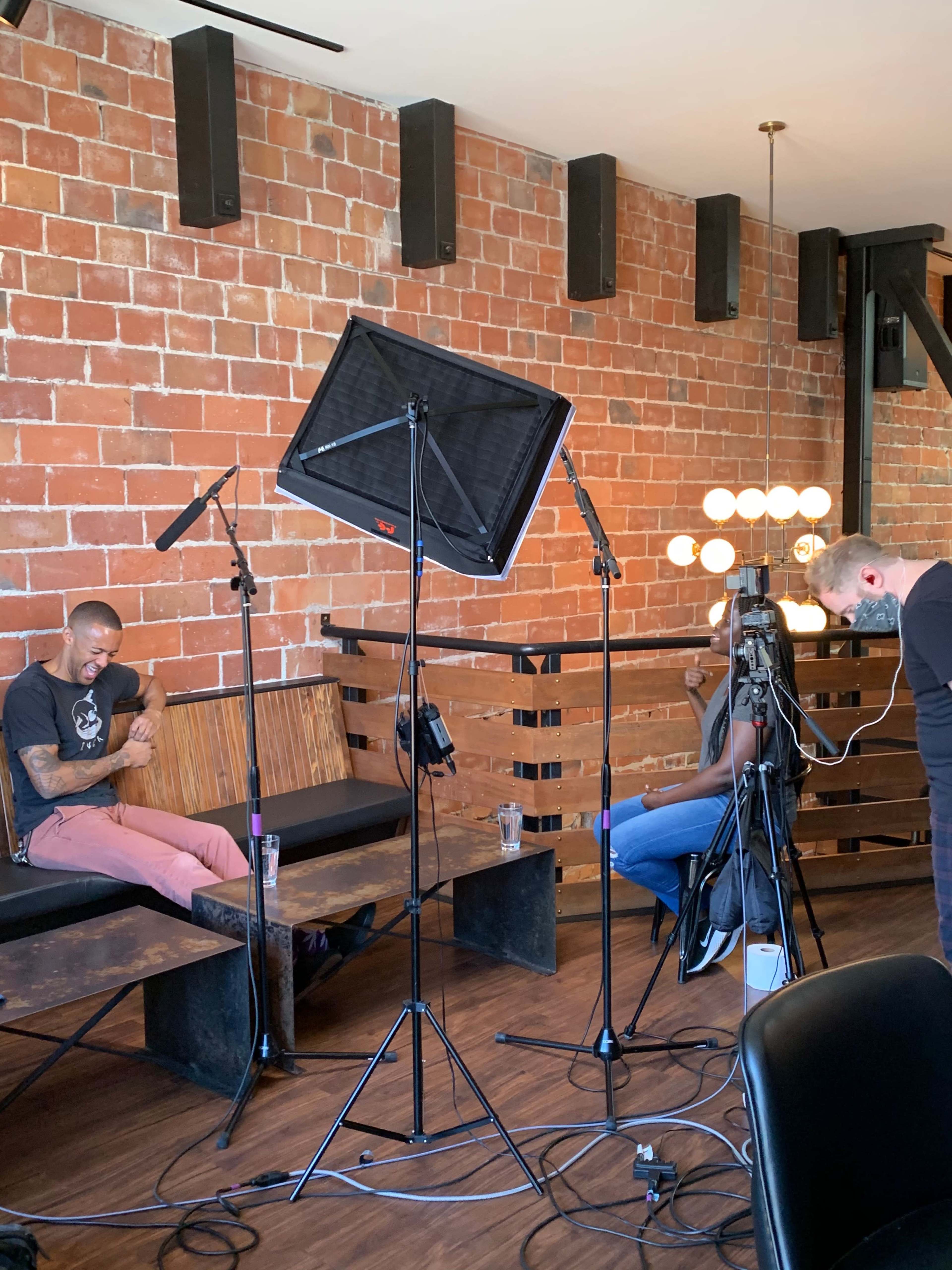 A man sits on a bench while another person operates a camera in a room with exposed brick walls and studio lighting equipment.