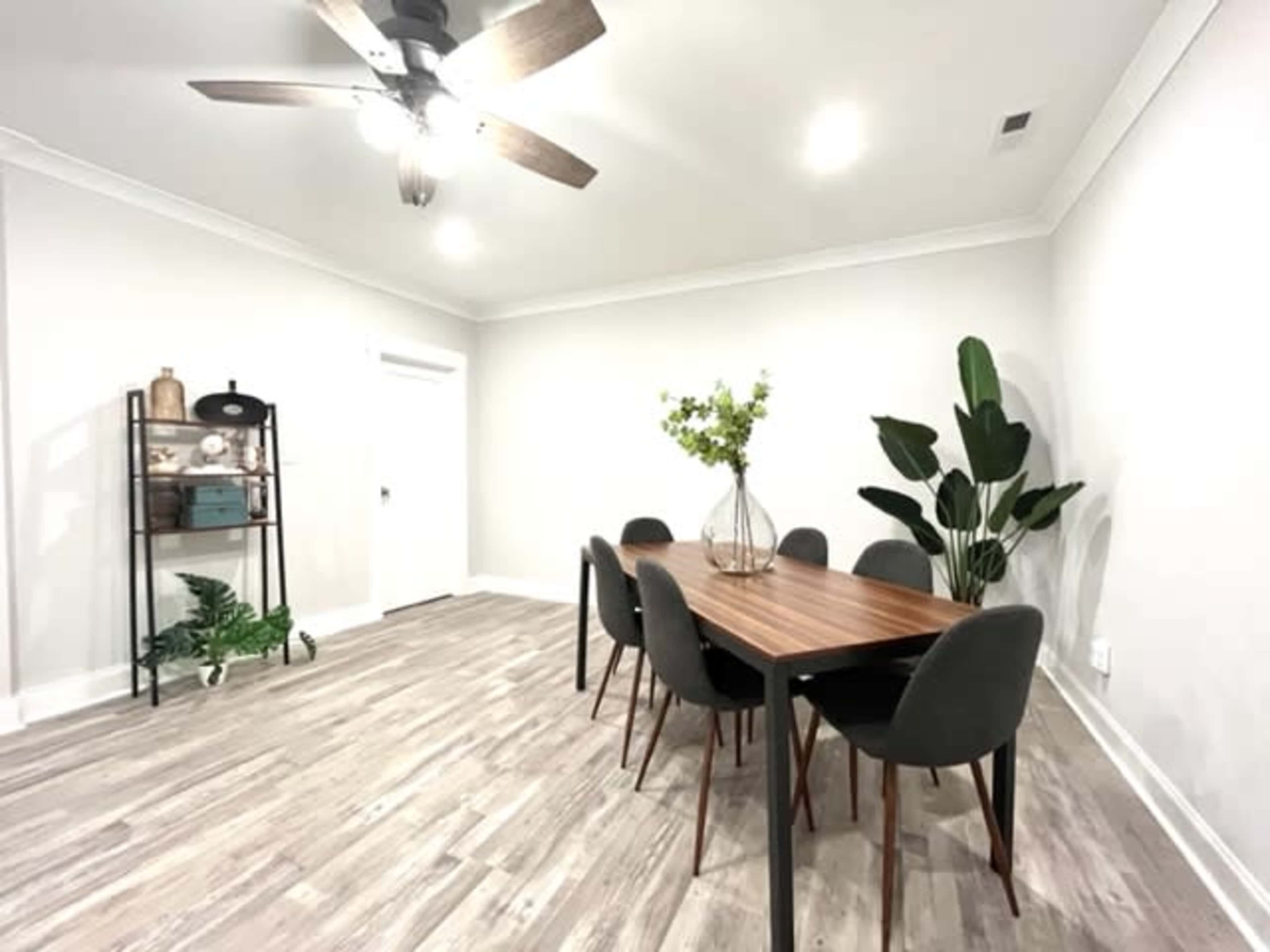 A modern dining room features a wooden table surrounded by six chairs, with a plant, a shelving unit, and light-colored walls.