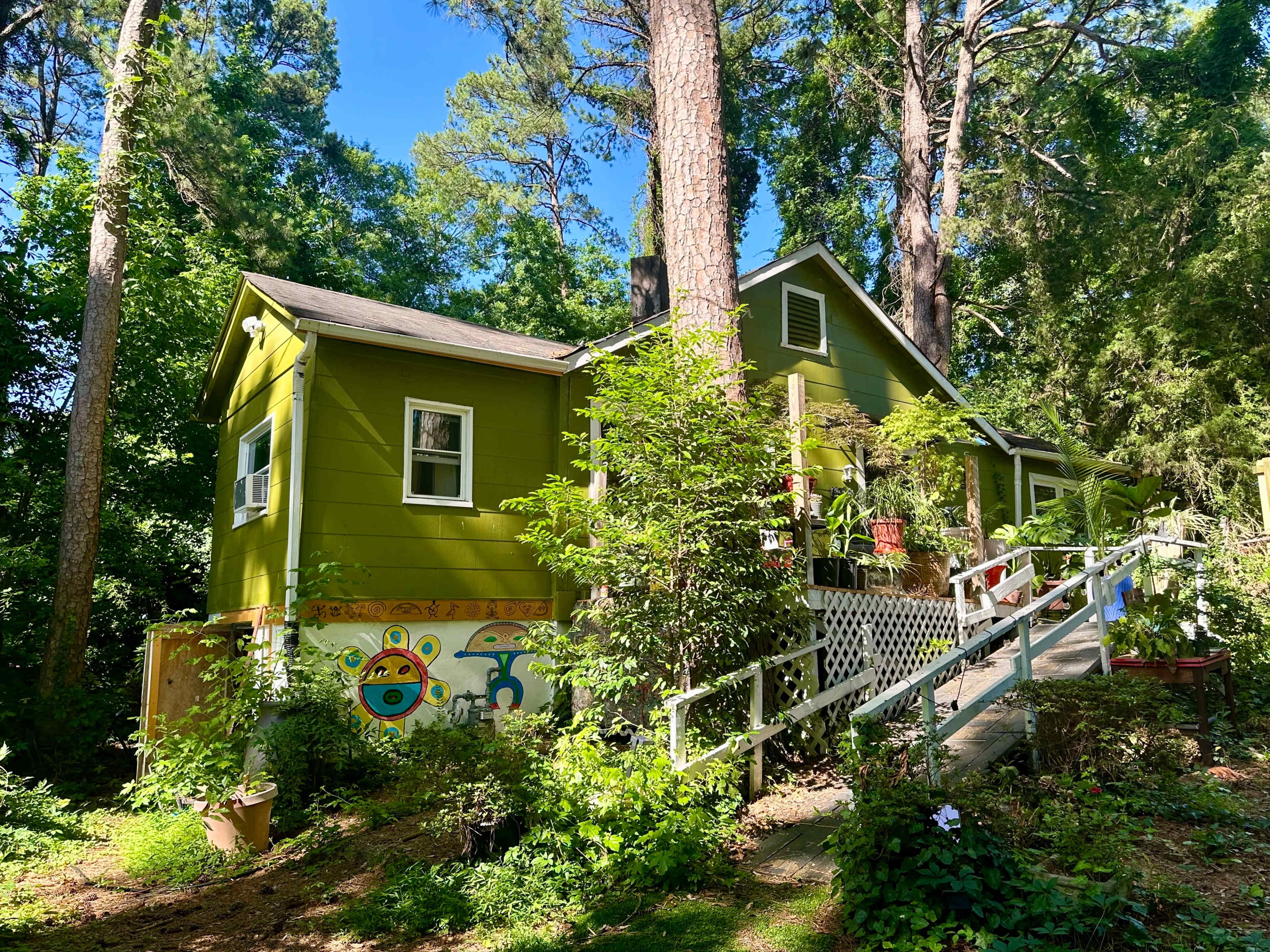 A green house with a decorative mural on one side is surrounded by trees and plants, featuring a front ramp leading to the entrance.