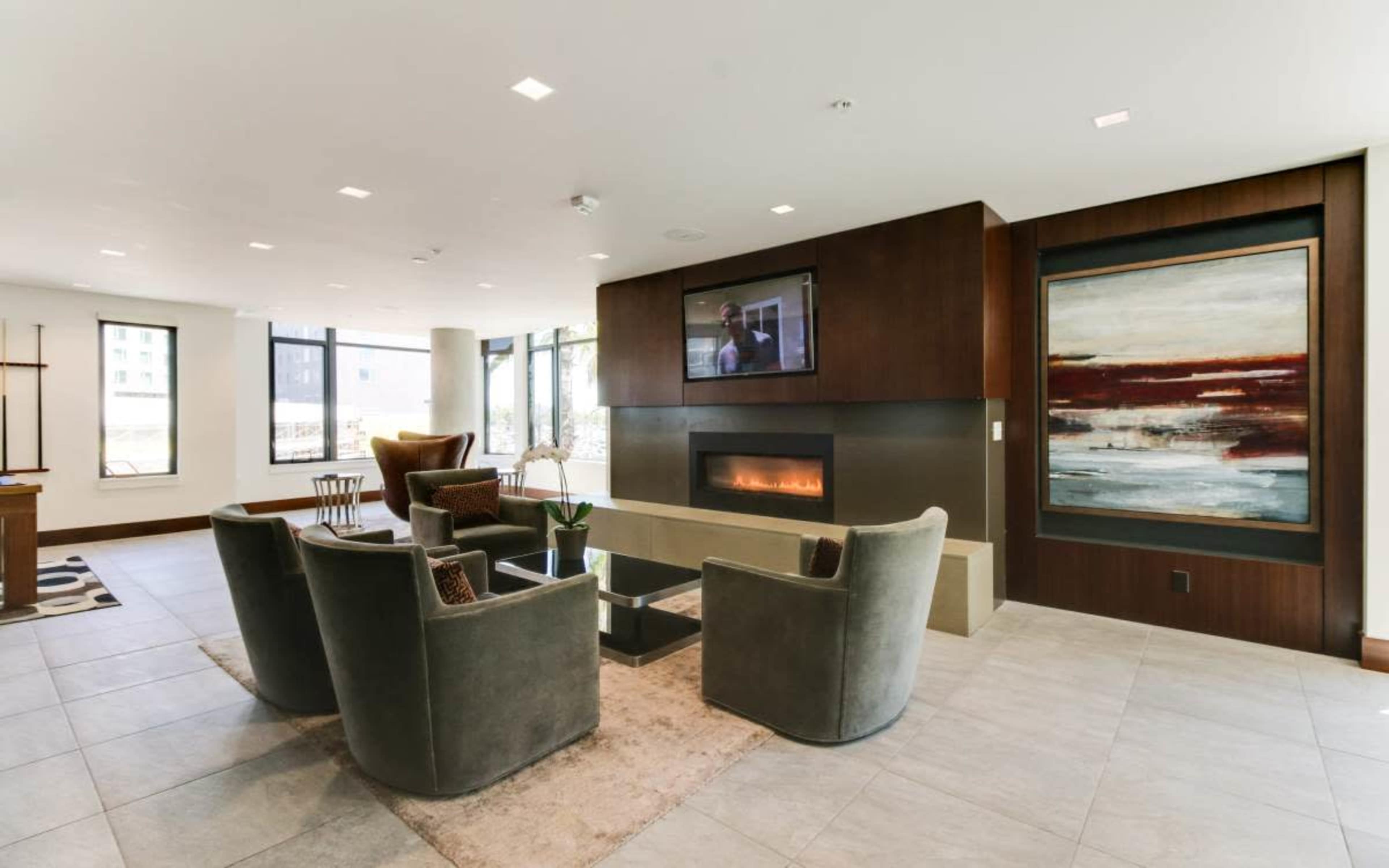 The image shows a modern living area featuring a fireplace, comfortable seating arranged around a coffee table, and large windows providing natural light.