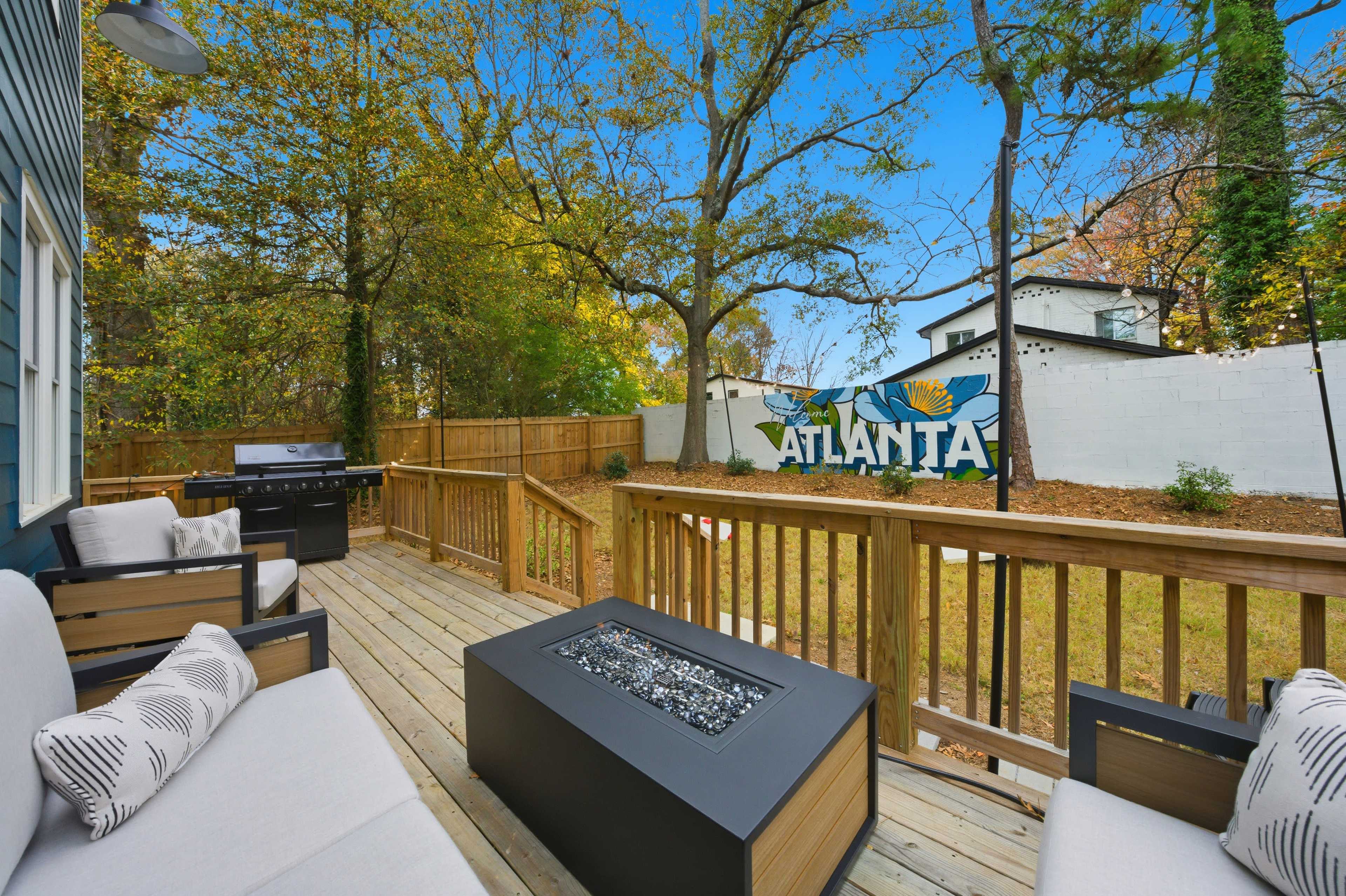 A wooden patio features outdoor seating and a fire pit, overlooking a mural that reads "ATLANTA" against a backdrop of trees and a white wall.