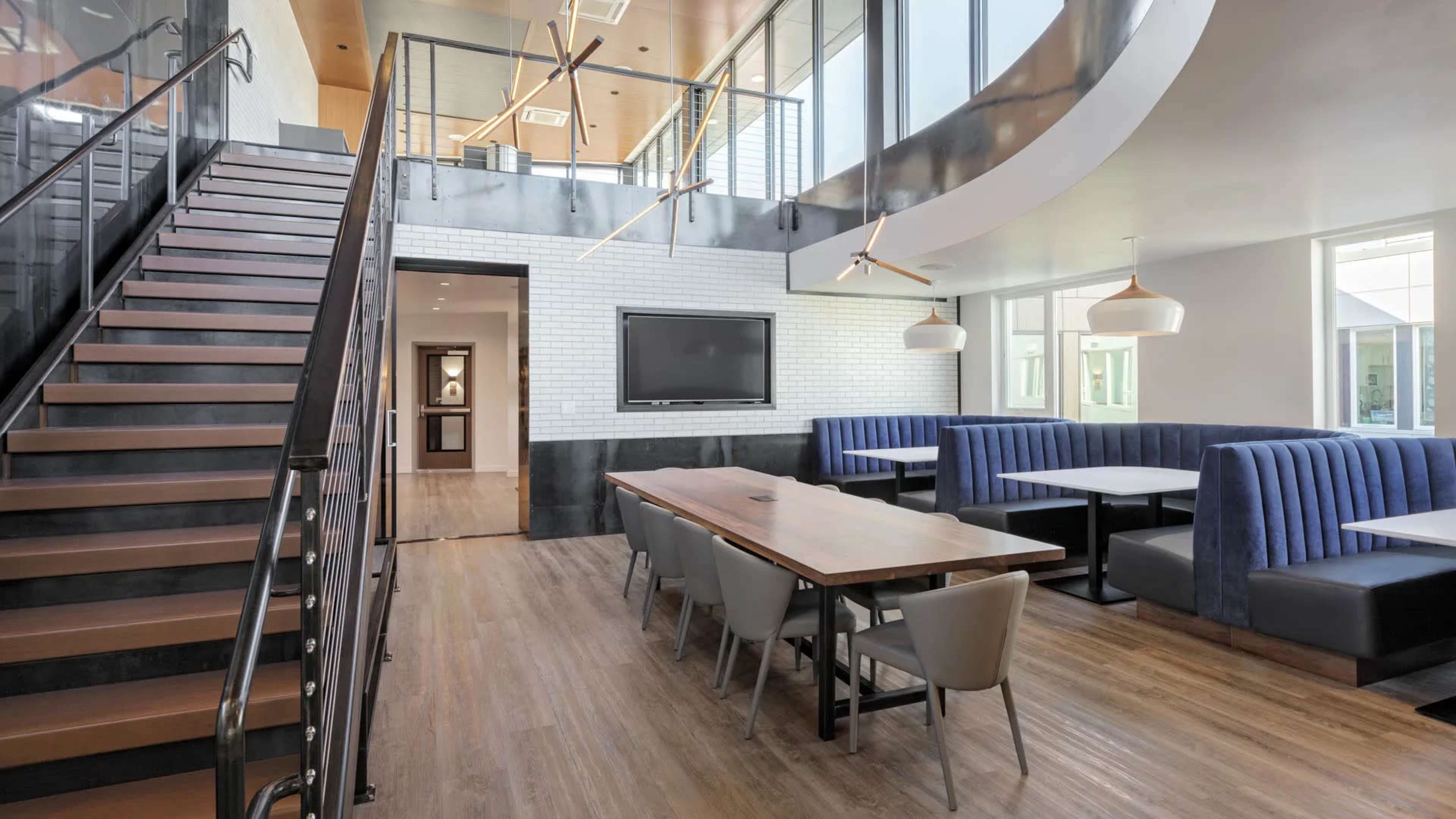 A modern interior space featuring a staircase, a large dining table surrounded by chairs, booth seating, and a flat-screen television on the wall.