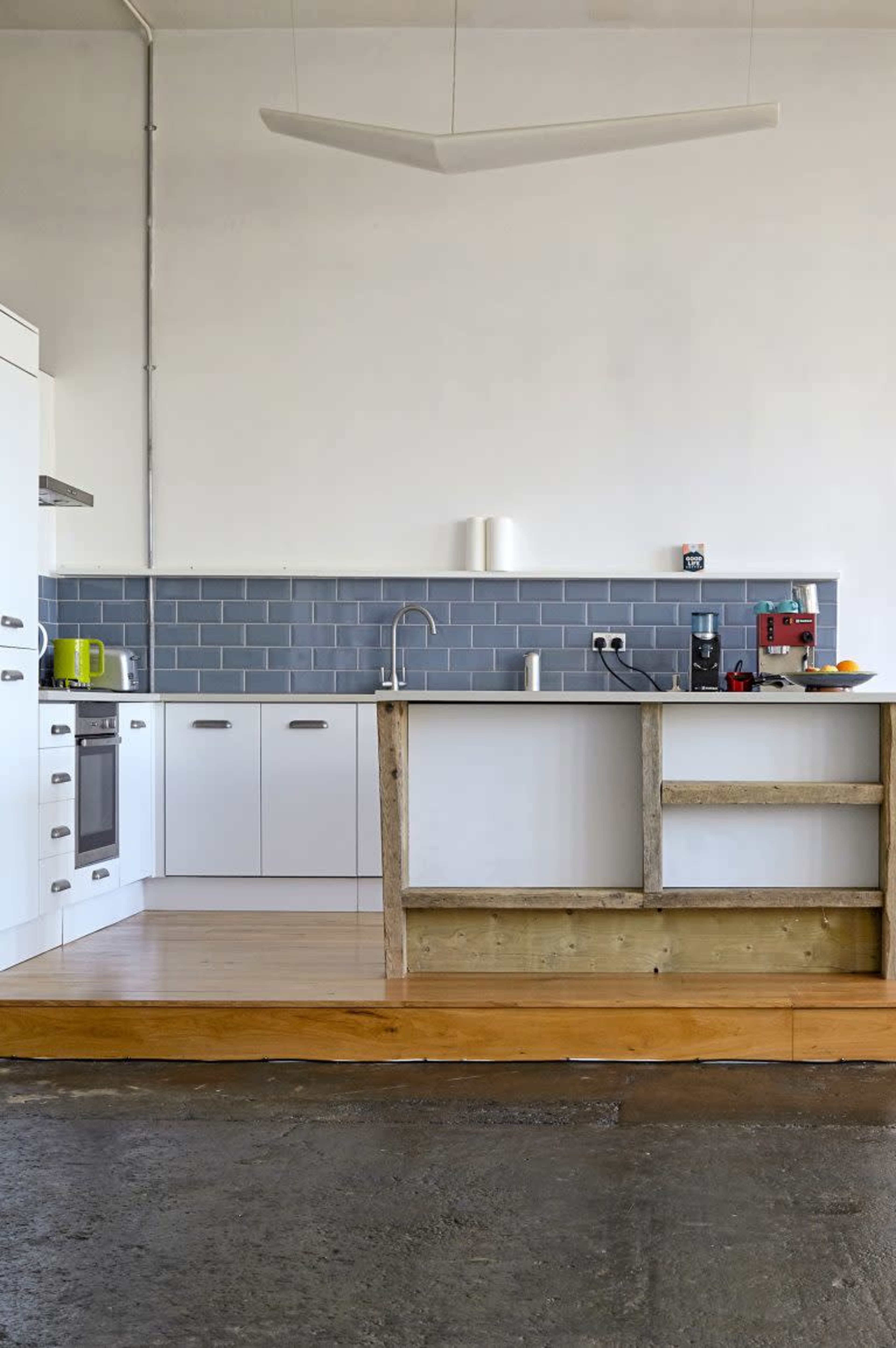 The image shows a modern kitchen with white cabinets, a gray tiled backsplash, and a wooden countertop on a raised platform.