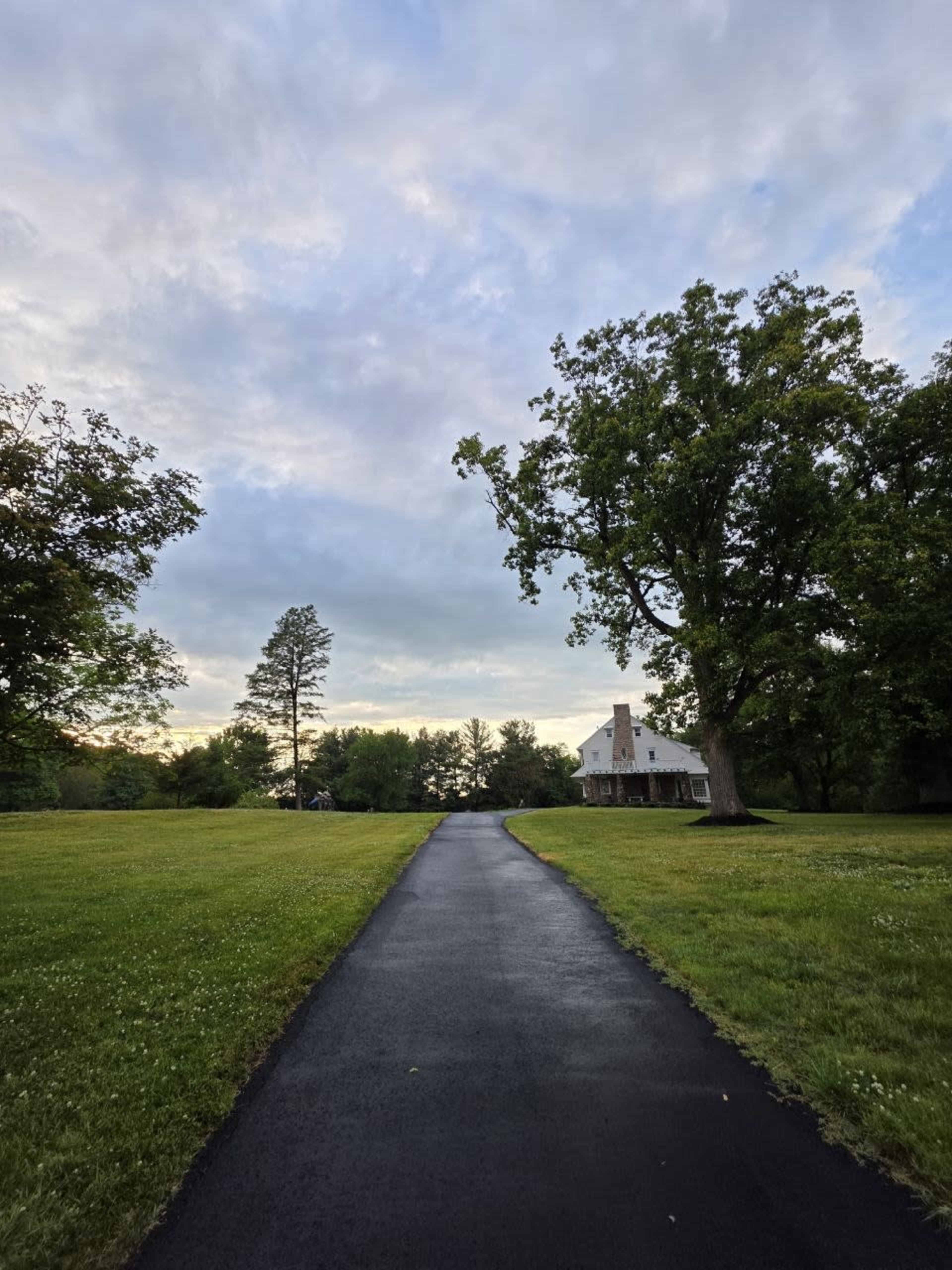 A paved pathway leads through a green lawn towards a house situated among trees under a cloudy sky.