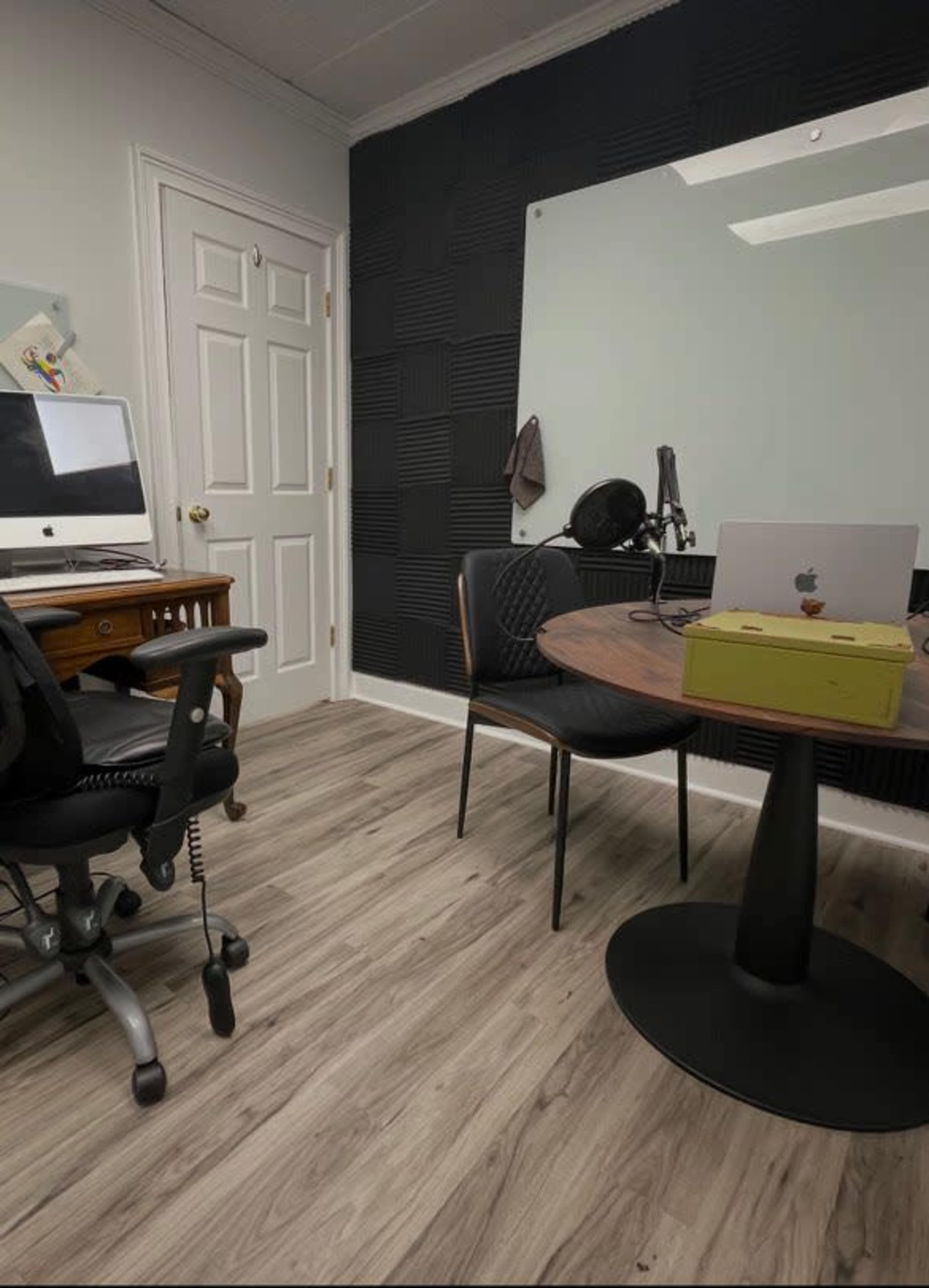 The image shows a small office space featuring a desk with a computer, a round table with a laptop and a box, and a wall equipped with sound paneling.