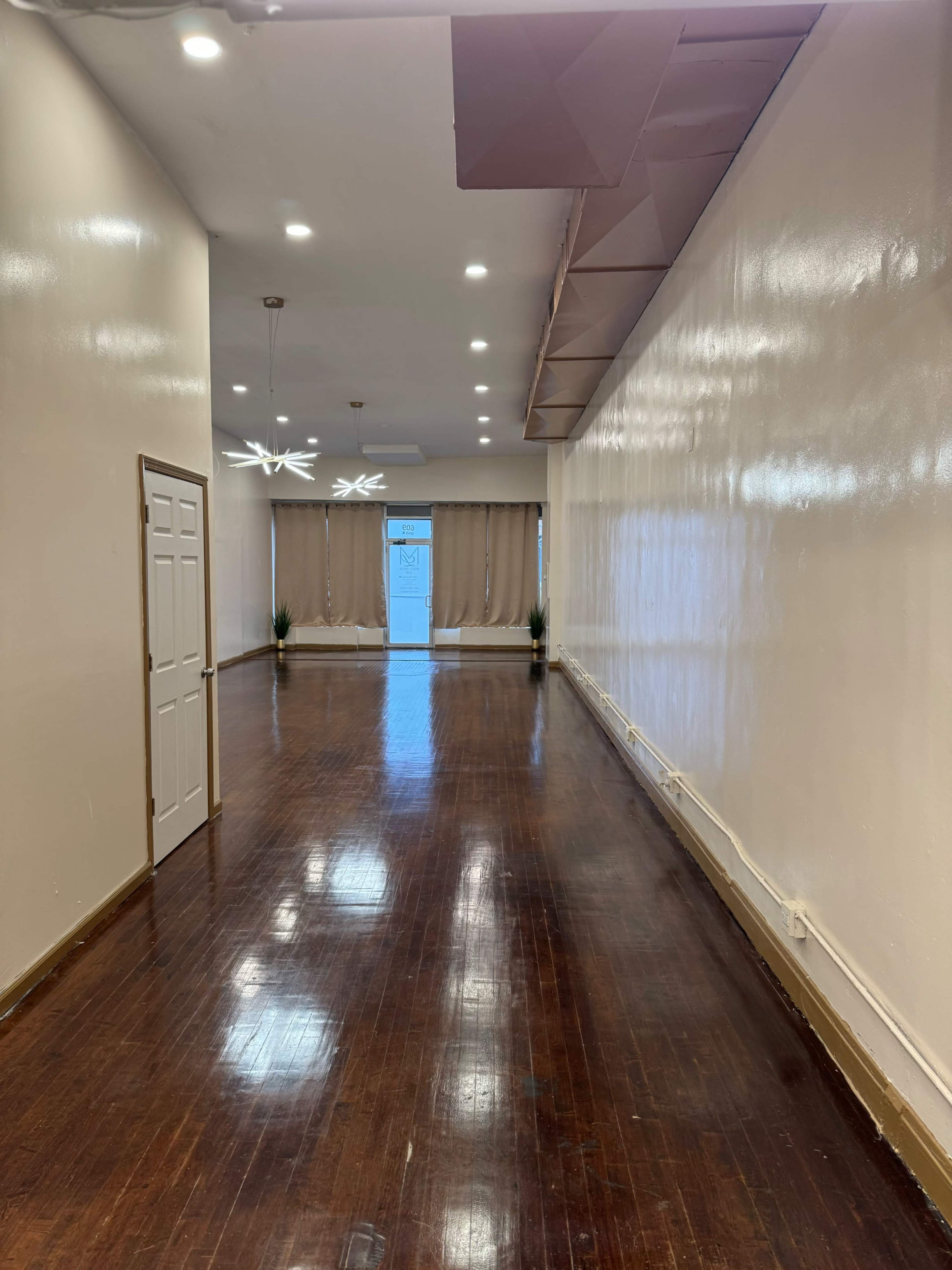 A well-lit, empty hallway with shiny wooden floors, leading to a door at the far end, flanked by two plants.
